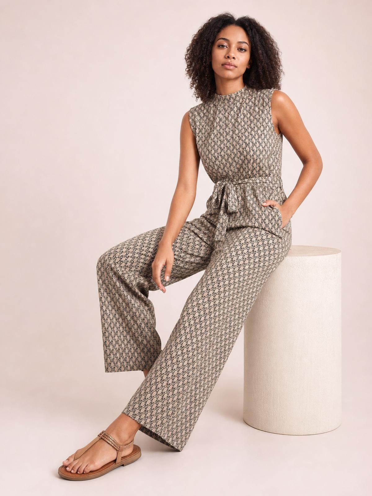 Model seated in a printed mock neck sleeveless jumpsuit with a tied waist, button detail, pockets, and wide-leg silhouette in taupe and black.