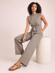 Model seated in a printed mock neck sleeveless jumpsuit with a tied waist, button detail, pockets, and wide-leg silhouette in taupe and black.