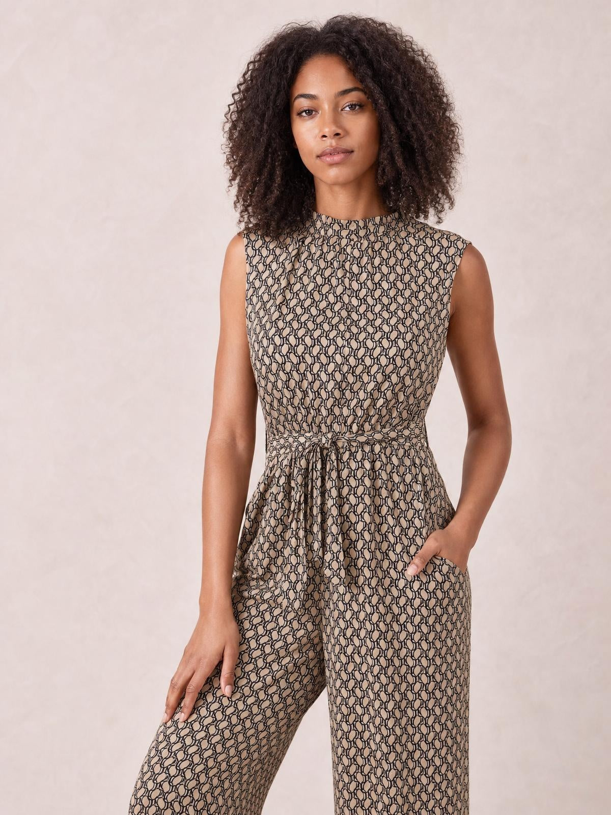 Model stands in a printed mock neck sleeveless jumpsuit with a tied waist, wide-leg silhouette, and side pockets, placing one hand in a pocket.