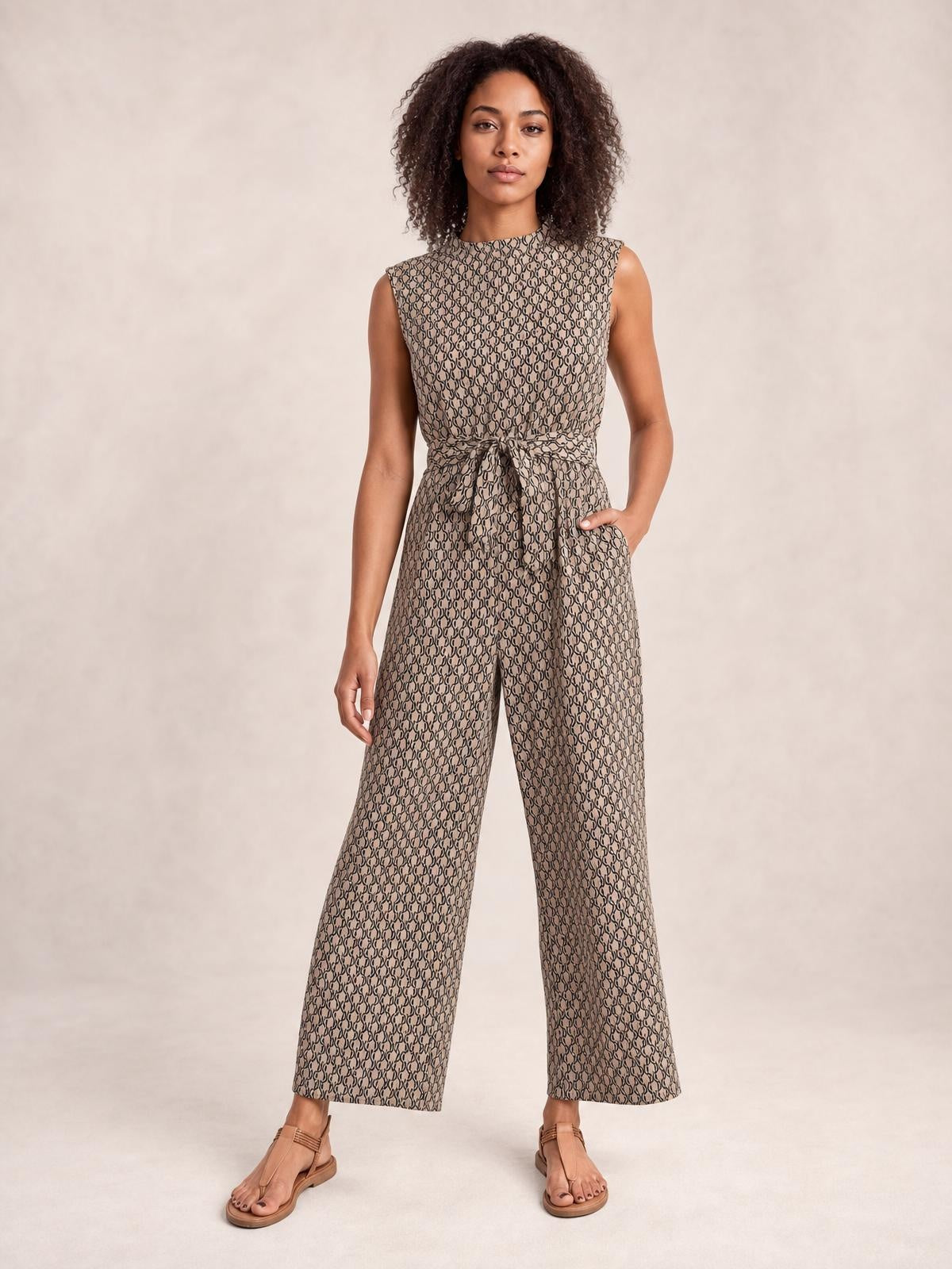 A model stands in a taupe printed mock neck sleeveless jumpsuit with a tied waist, side pockets, and a relaxed wide-leg silhouette.