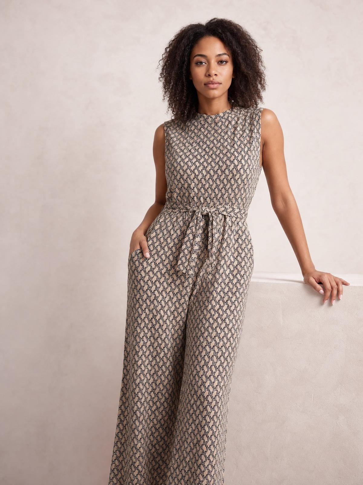 Model poses in a beige printed mock neck sleeveless jumpsuit with a tied waist, wide-leg silhouette, and visible side pockets.