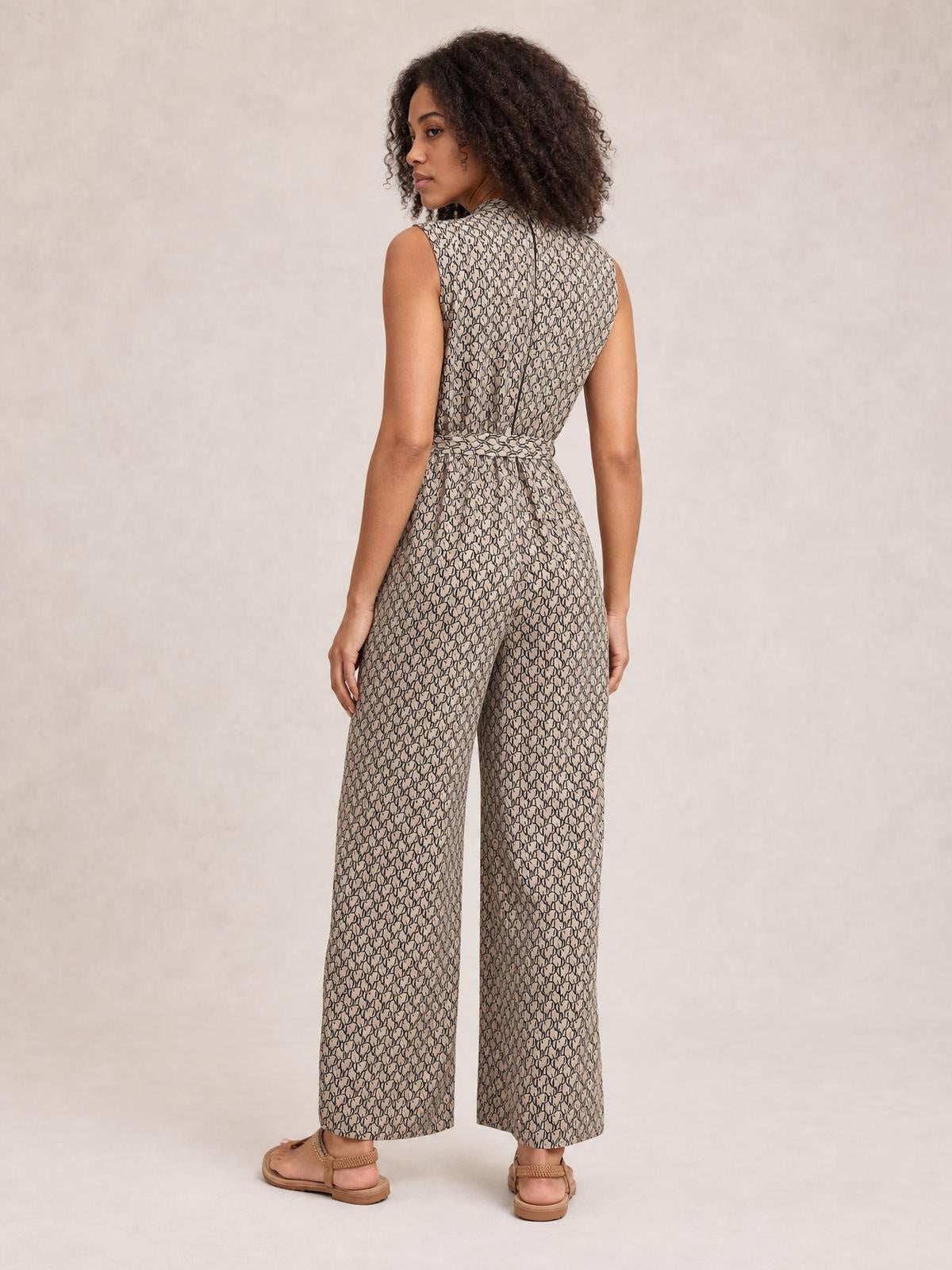 Model shown from behind wears a beige-and-black printed sleeveless mock neck jumpsuit with a tied waist and flowing wide-leg silhouette.