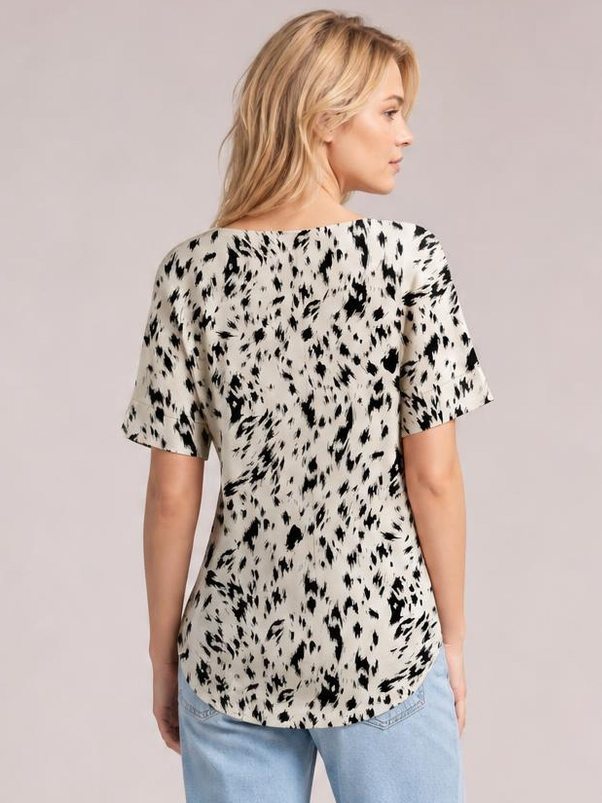 Model turned to show the back of a cream printed blouse with black abstract spots, regular half sleeves, a relaxed regular silhouette, and curved hem.