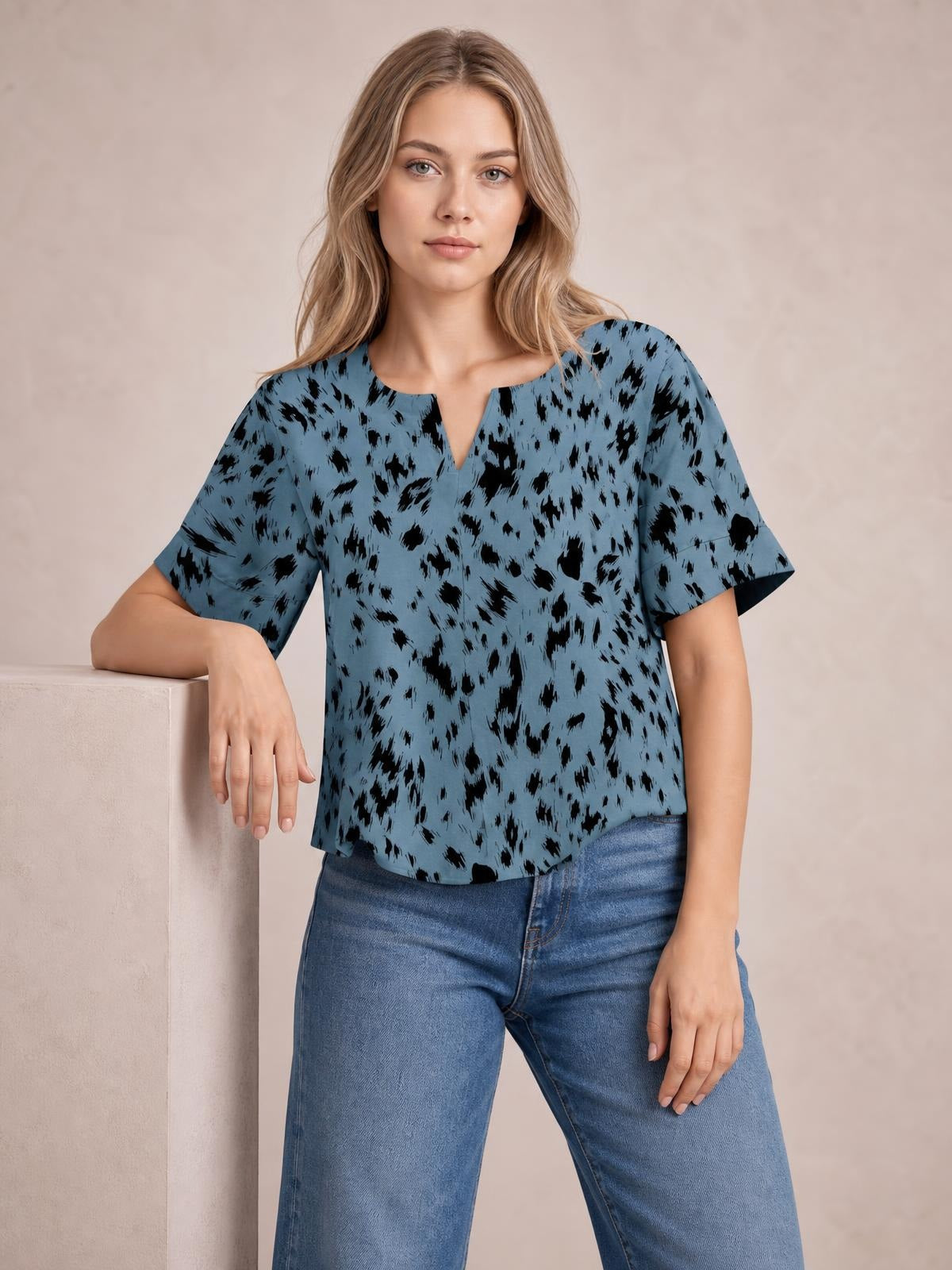 Model wearing a blue printed blouse with black abstract spots, notched neckline, regular fit, and half sleeves, posing with one hand resting on a ledge.