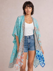 Model wearing a turquoise printed open front cover-up with a flowing, loose silhouette and draped three-quarter sleeves over a white tank and denim shorts.