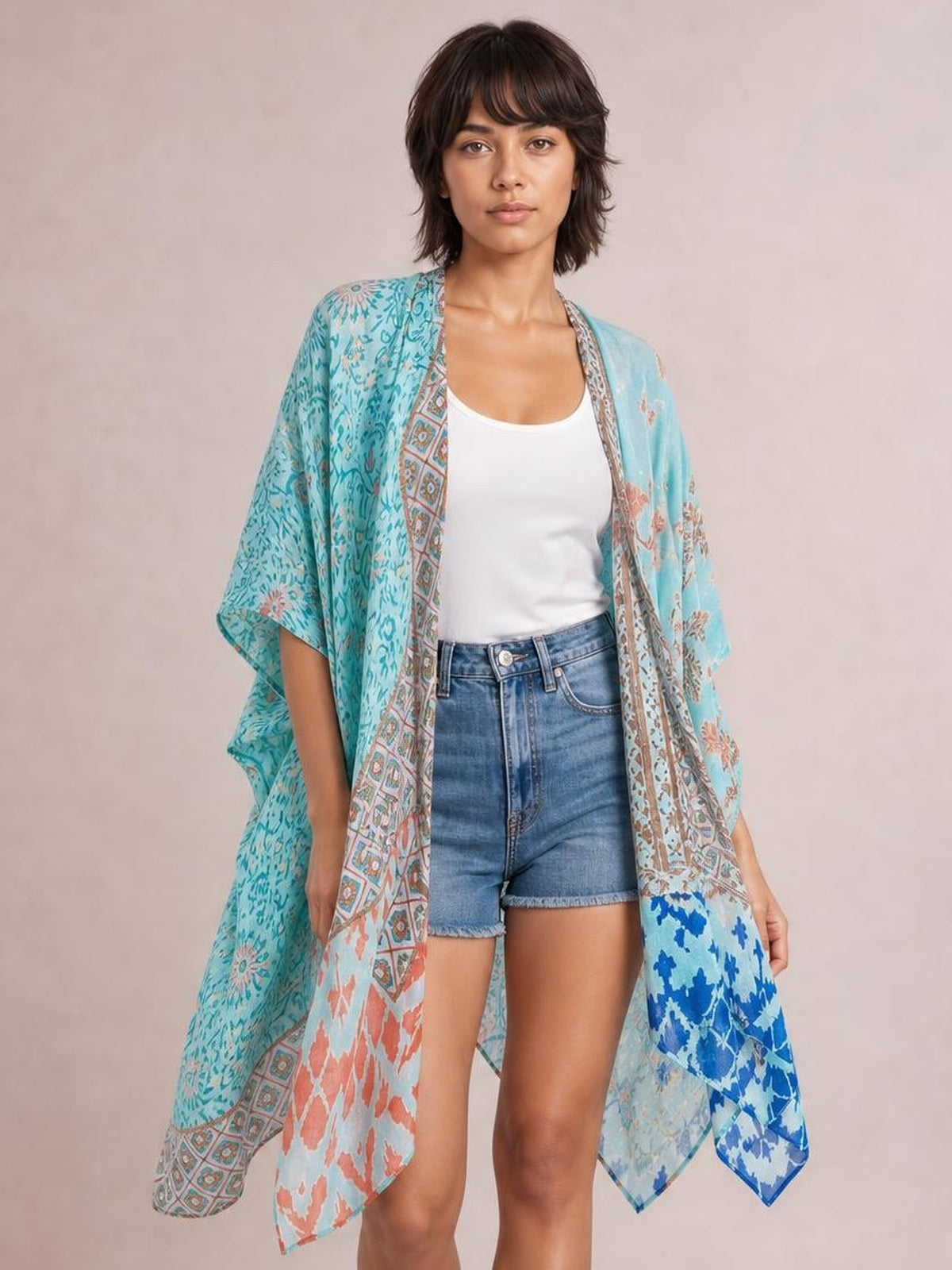 Model wearing a turquoise printed open front cover-up with a flowing, loose silhouette and draped three-quarter sleeves over a white tank and denim shorts.