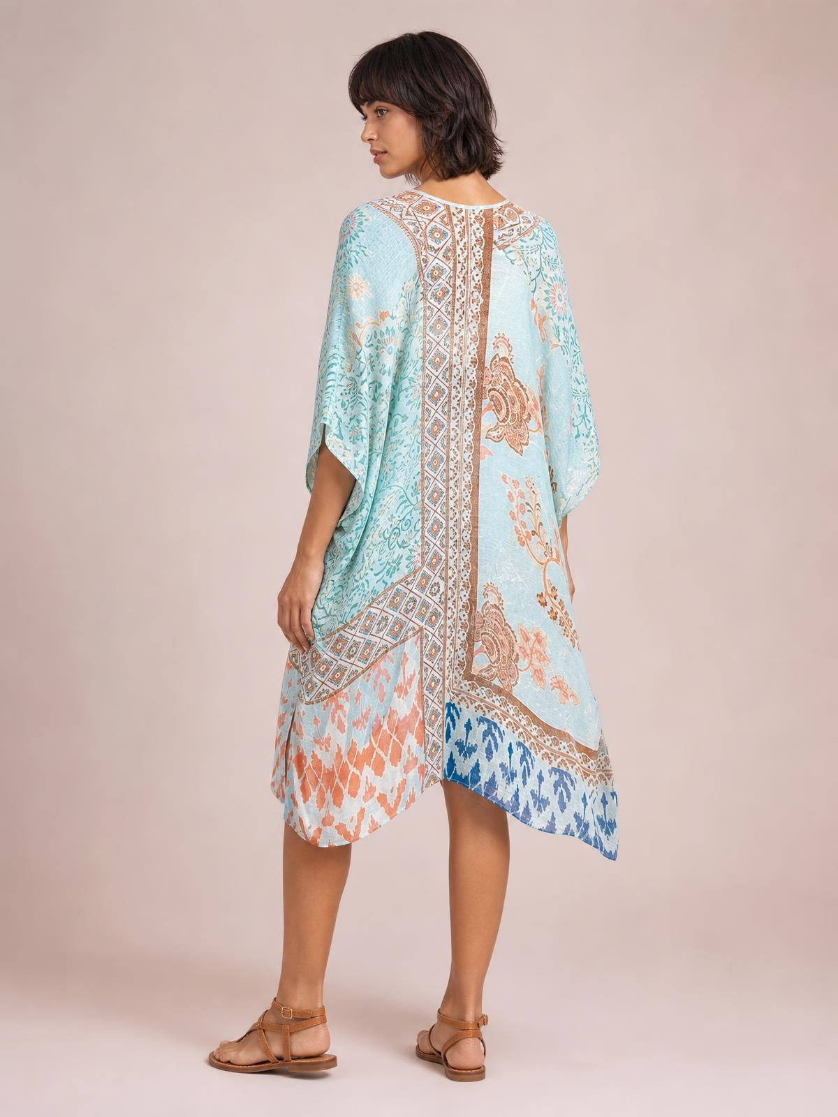 Model turned away in a relaxed pose wearing a light blue printed open front cover-up with flowing dolman sleeves, an oversized silhouette, and a curved hem.