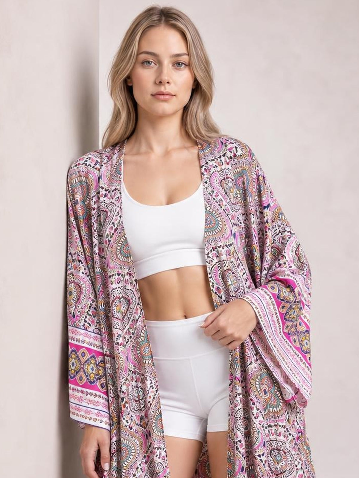 Model wears a pink and multicolor printed open front cover up with long sleeves, a relaxed draped silhouette, and one hand resting at her waist.