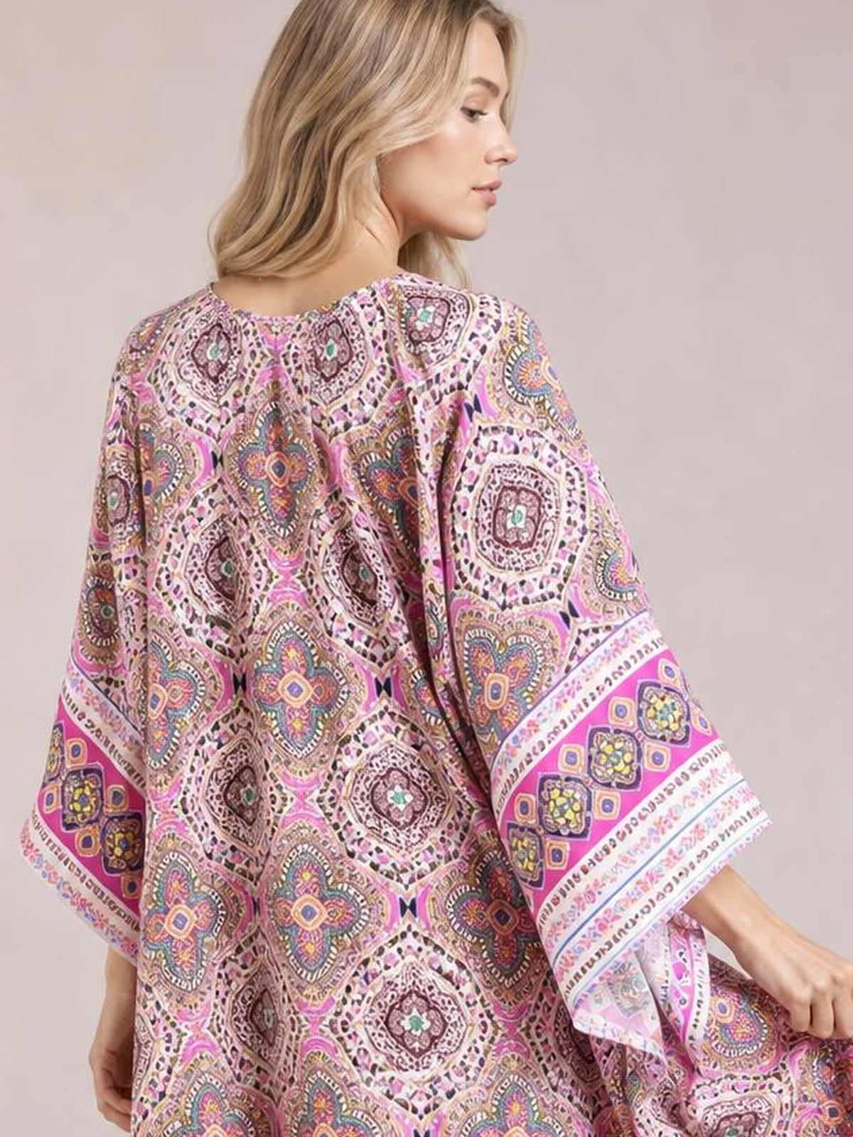 A model turned slightly away wears a flowing pink printed cover up with a loose silhouette, wide sleeves, and decorative patterned borders.