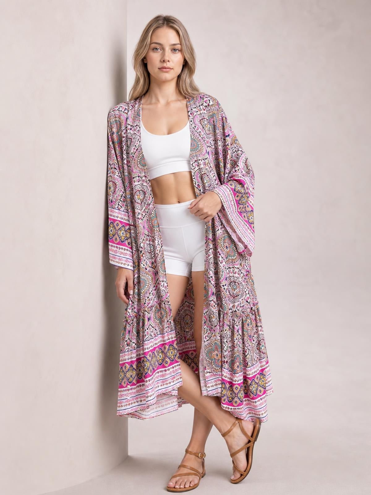 A model wears a long, flowing printed open-front cover up in pink, white, and multicolor paisley, with long sleeves and a tied closure over white separates.