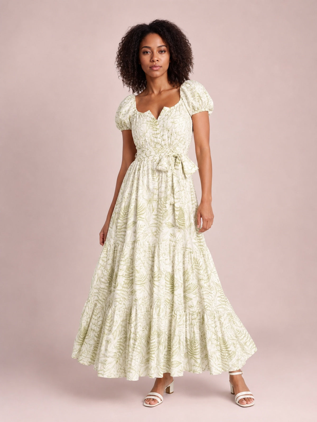 A model stands in a flowing botanical-print maxi dress with a ruched bodice, short puff sleeves, tied waist bow, and tiered full-length silhouette.