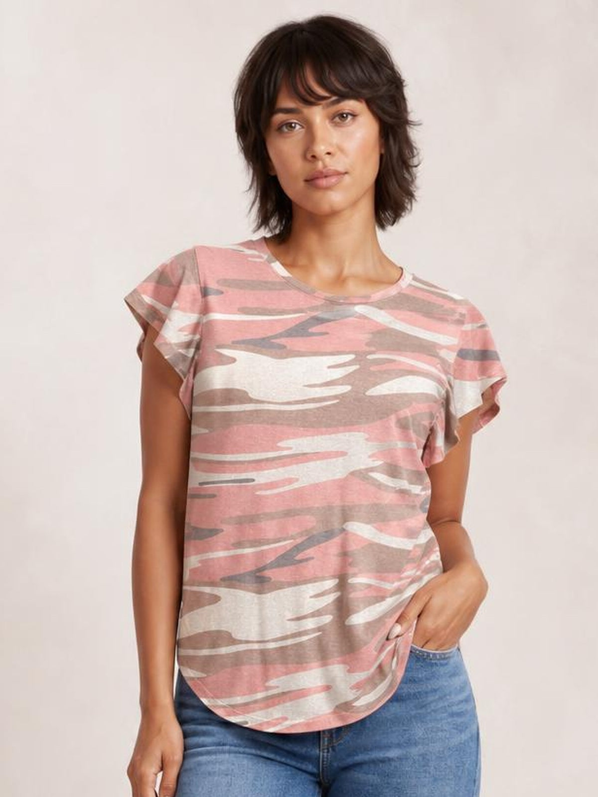 Model wearing a pink, taupe, and white printed round-neck short-sleeve T-shirt with a relaxed silhouette and curved hem, posing with one hand in her jeans pocket.