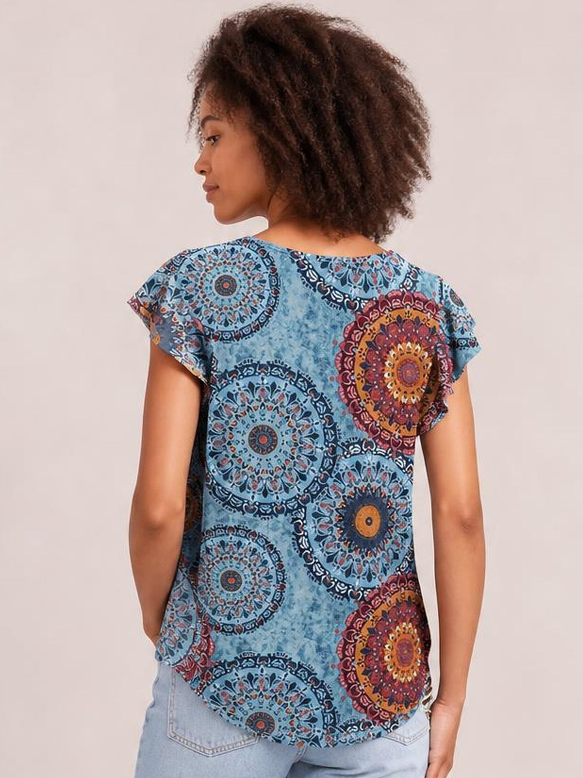 A model looks over her shoulder wearing a blue printed round neck short sleeve T-shirt with ornate multicolor circular motifs, a relaxed silhouette, and curved hem.