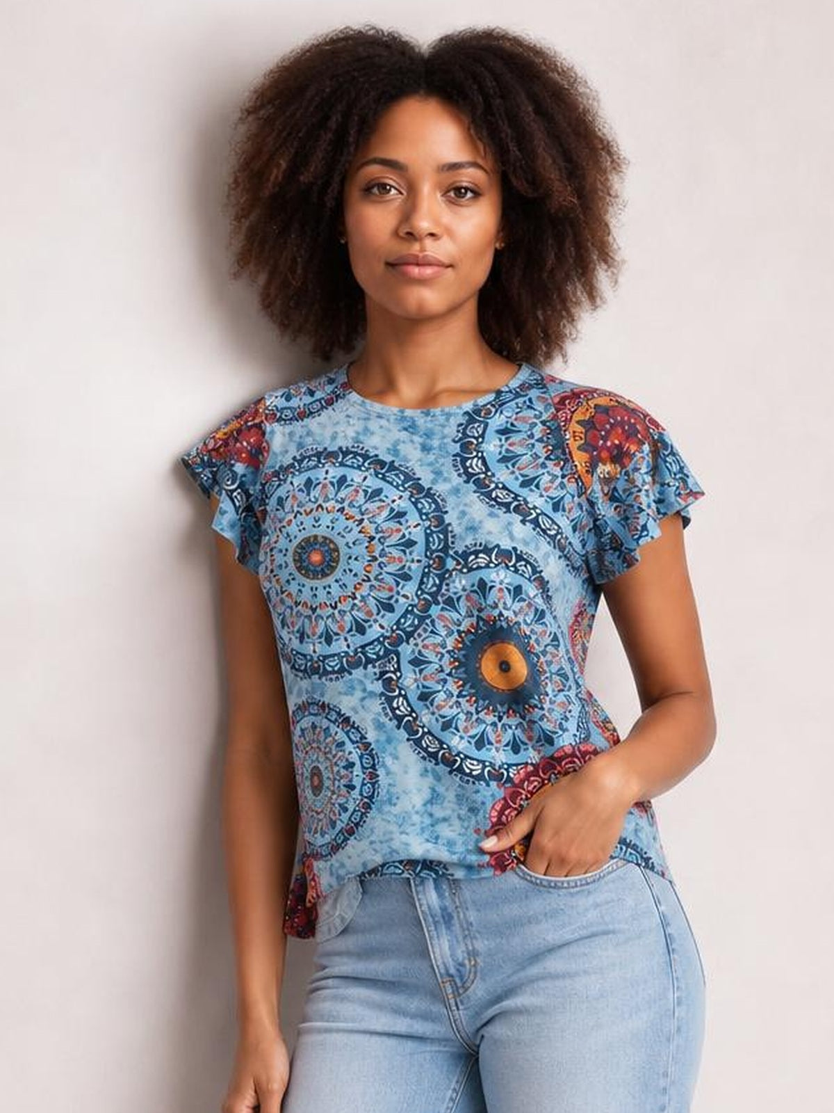 A model wears a blue printed round-neck short-sleeve T-shirt with a relaxed silhouette and colorful medallion pattern, posing with one hand in her jeans pocket.