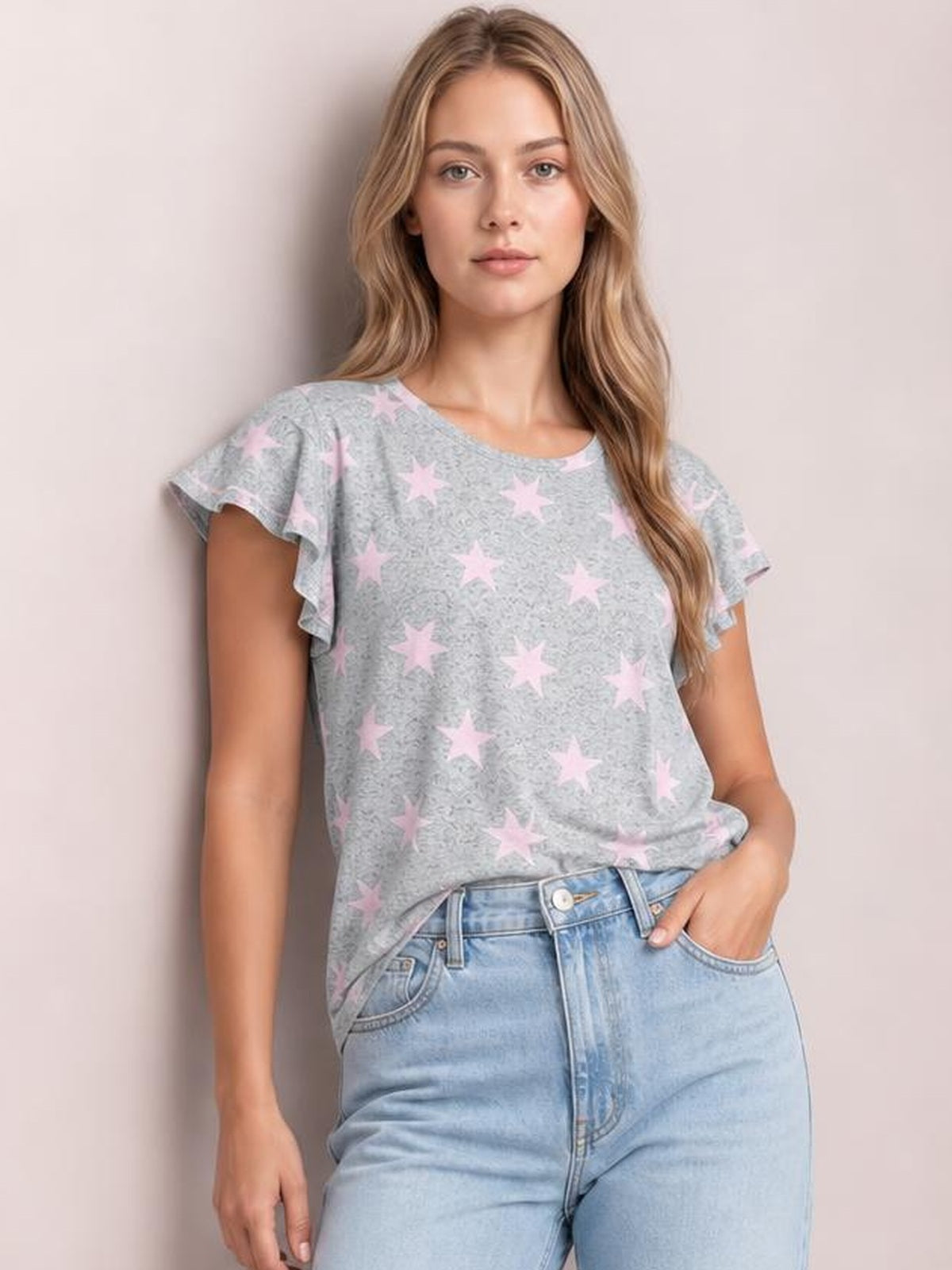 A model poses in a gray printed round neck short sleeve T-shirt with pink stars, ruffled sleeve edges, and a relaxed silhouette, one hand in her jeans pocket.