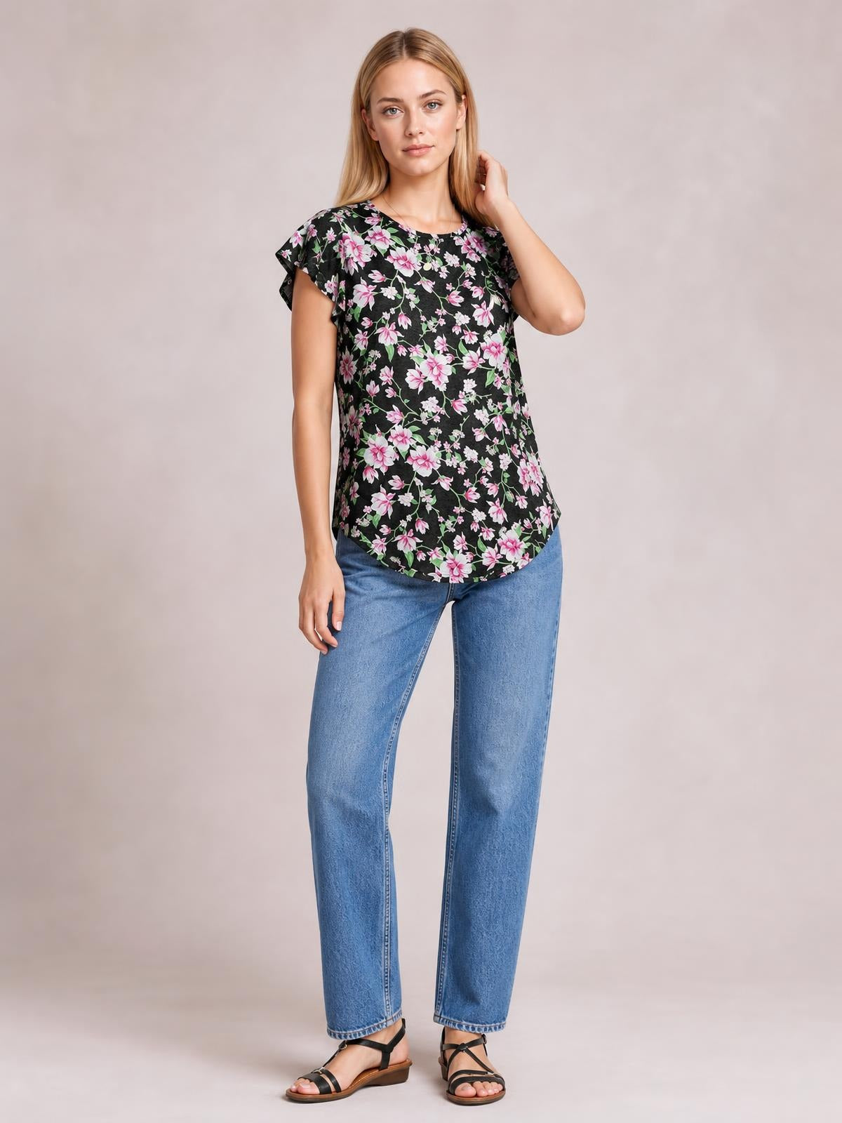 A woman poses in a black floral printed round neck short sleeve T-shirt with flutter sleeves, a relaxed silhouette, and a curved hem.