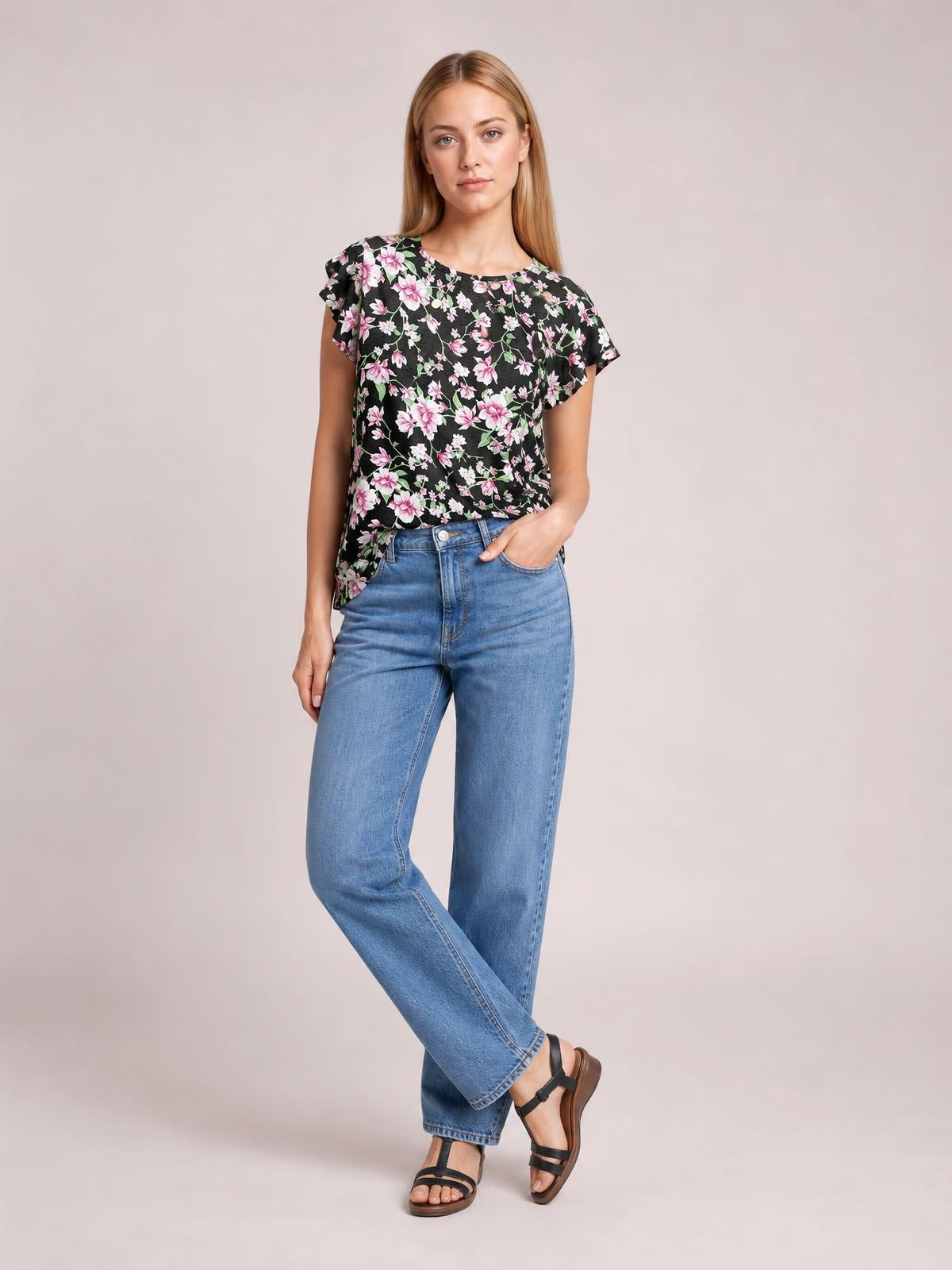 Model wears a black printed round neck short sleeve T-shirt with a pink floral pattern and relaxed silhouette, standing with crossed legs and one hand in her pocket.