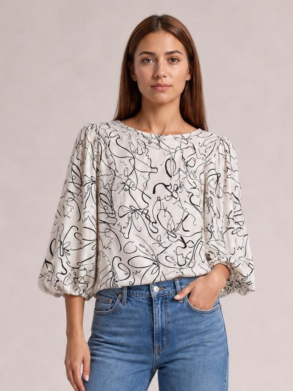 Model wears an ivory printed round-neck blouse with black abstract floral lines, a relaxed silhouette, and gathered three-quarter sleeves, posing with one hand in her jeans pocket.