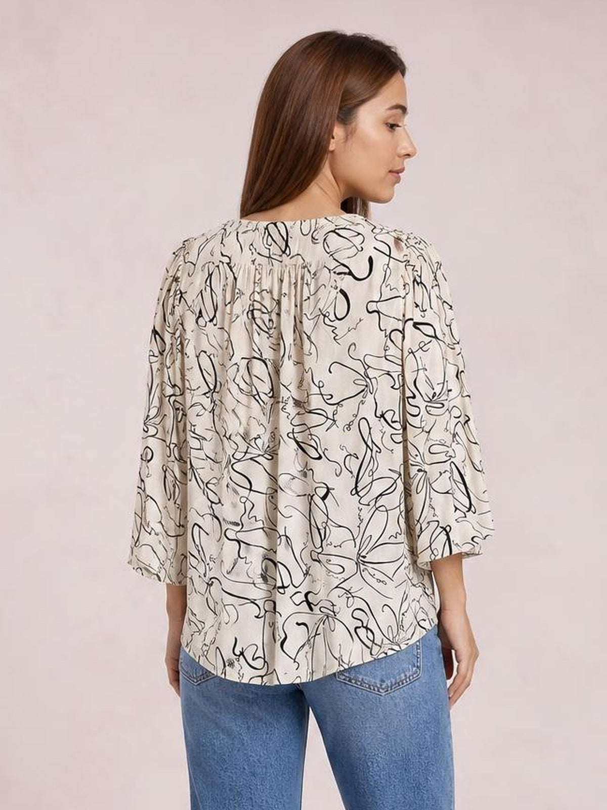 Model shown from the back in a cream printed blouse with a round neck, three-quarter sleeves, relaxed silhouette, and gathered back yoke.