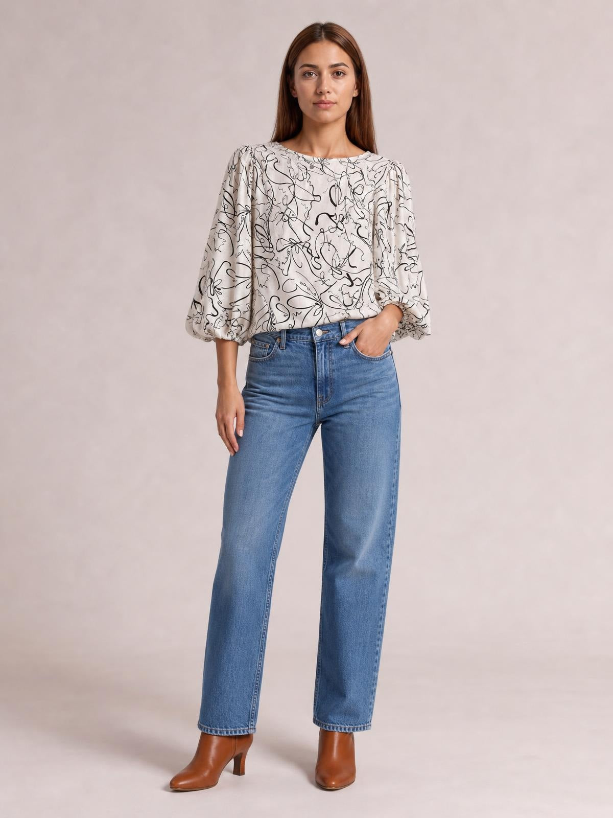 Model wears a cream printed round neck blouse with black abstract lines, voluminous three-quarter sleeves, and a relaxed silhouette, posing with one hand in her jeans pocket.