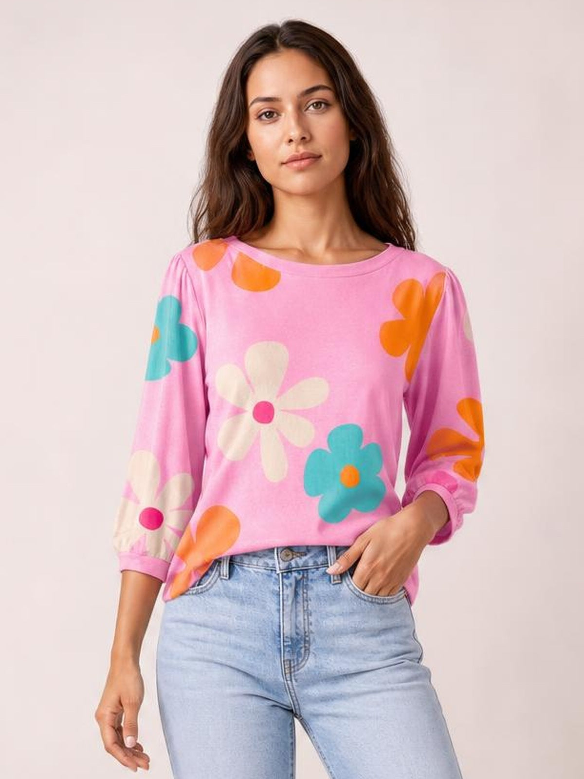 Model wears a pink printed round neck top with colorful floral motifs, long sleeves, and a relaxed silhouette, posing with one hand in her jeans pocket.