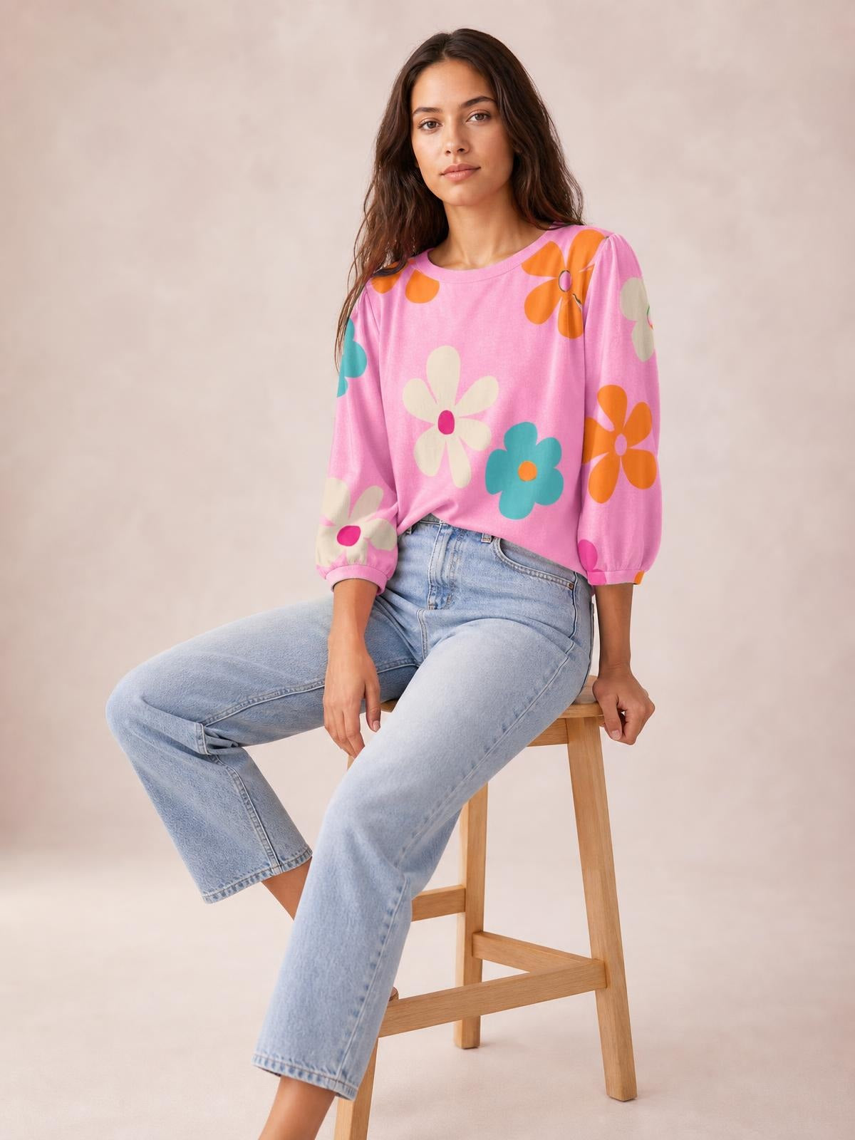 Model seated in light jeans wears a pink printed round neck top with colorful floral motifs, relaxed silhouette, and puffed three-quarter sleeves with gathered cuffs.