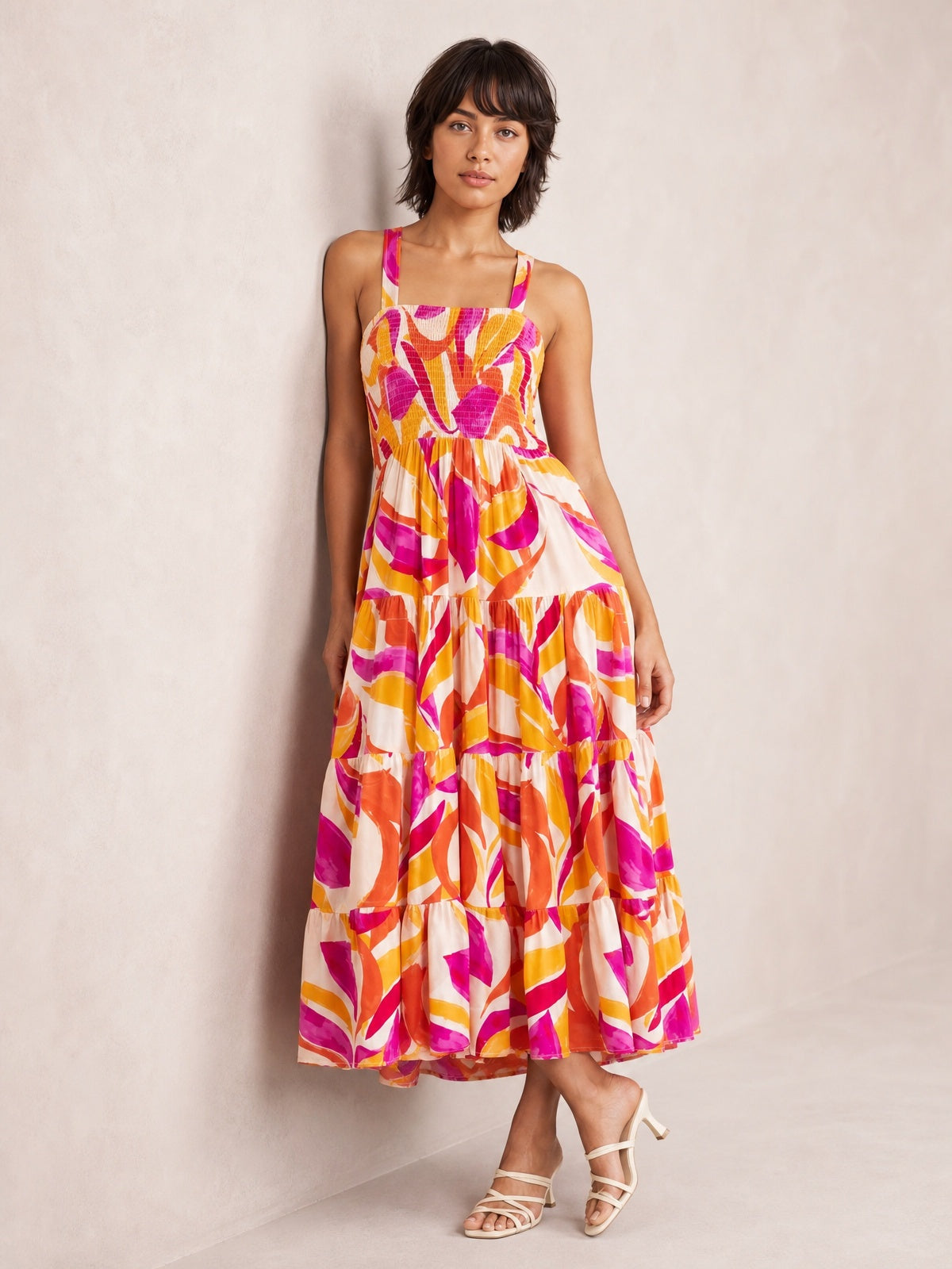 Model poses in a vibrant multicolored printed midi dress with a smocked square neckline, sleeveless cut, fitted bodice, and tiered A-line skirt.