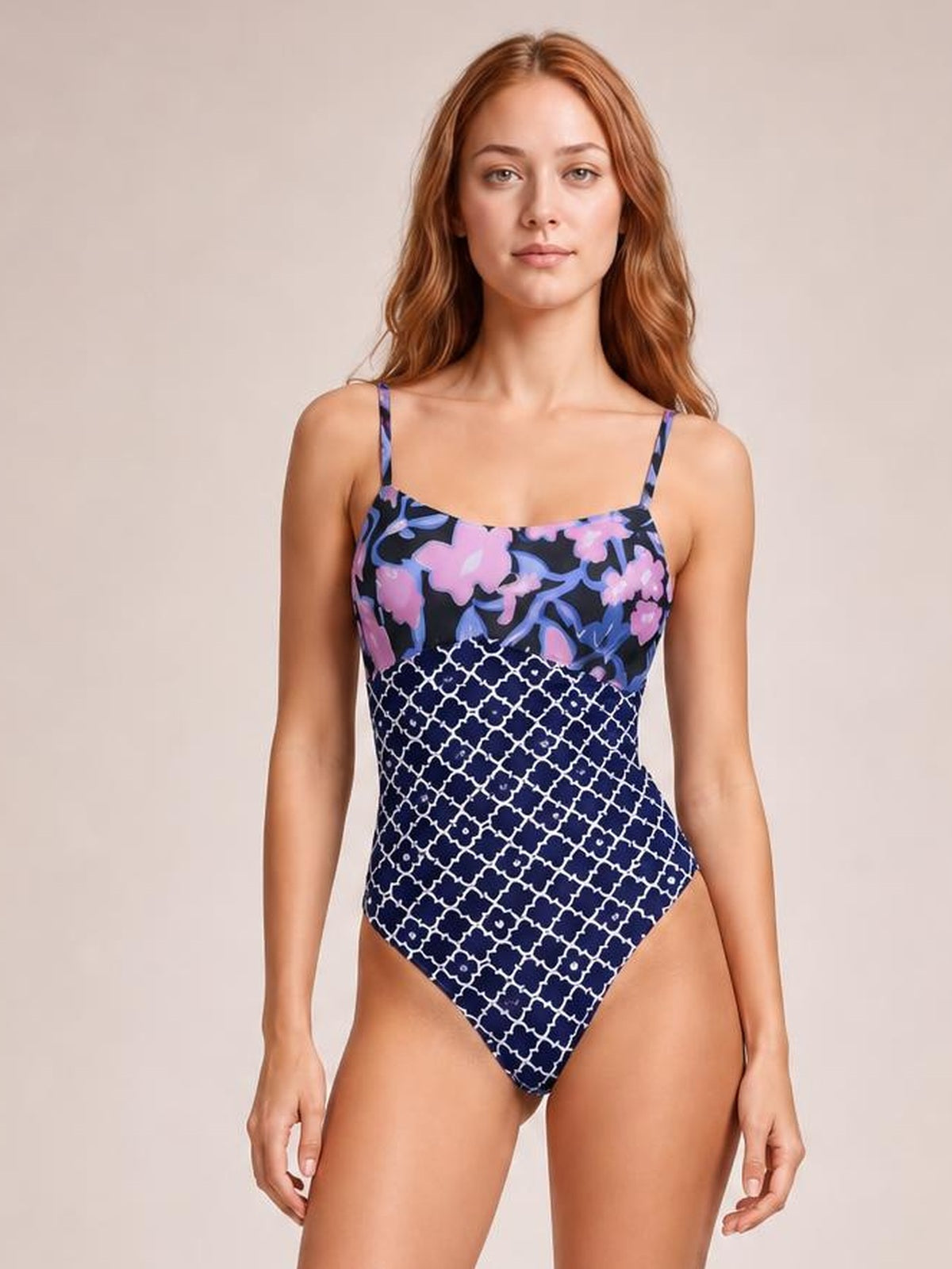 Front-facing model stands with arms relaxed in a printed spaghetti strap one-piece swimsuit with a sweetheart neckline, floral navy bodice, and white lattice-patterned lower panel.