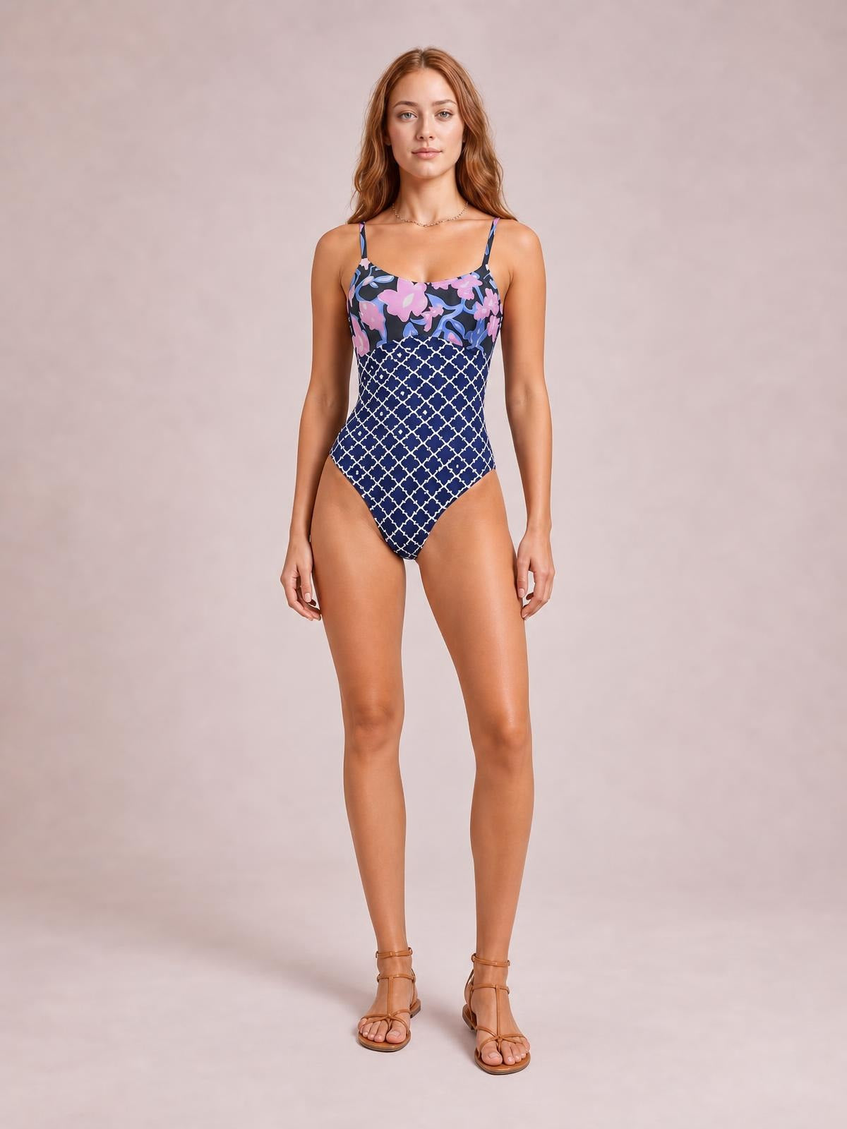 Front-facing model wears a stretchy printed one-piece swimsuit with thin spaghetti straps, sweetheart neckline, floral bodice, navy geometric pattern, and high-cut silhouette.