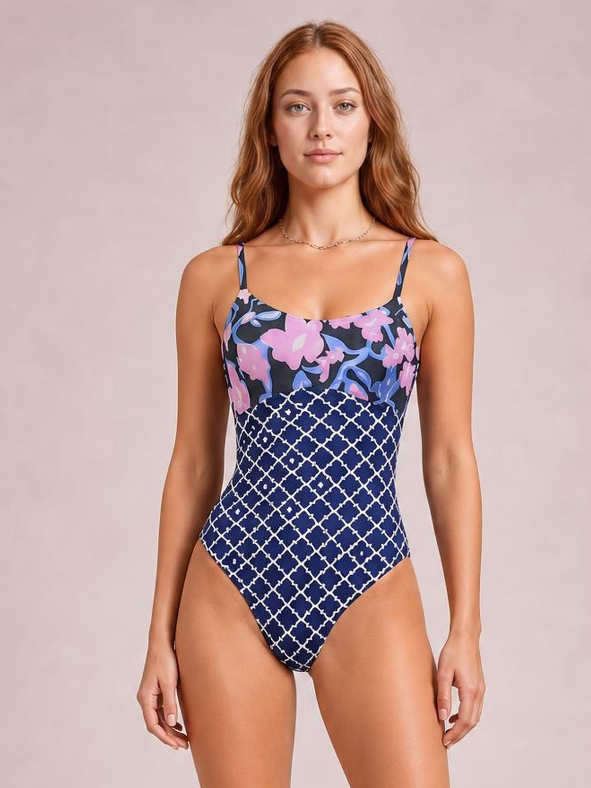 A model stands front-facing in a fitted printed one-piece swimsuit with spaghetti straps, a sweetheart neckline, floral bodice, and navy geometric-patterned lower section.
