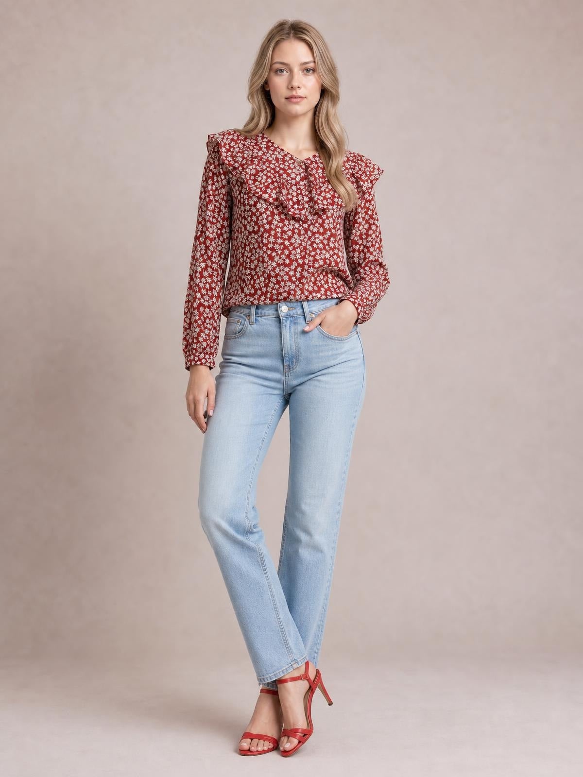 A model wears a red floral-print blouse with long sleeves, a ruffled statement collar, and a relaxed silhouette, standing with crossed legs and one hand in her pocket.