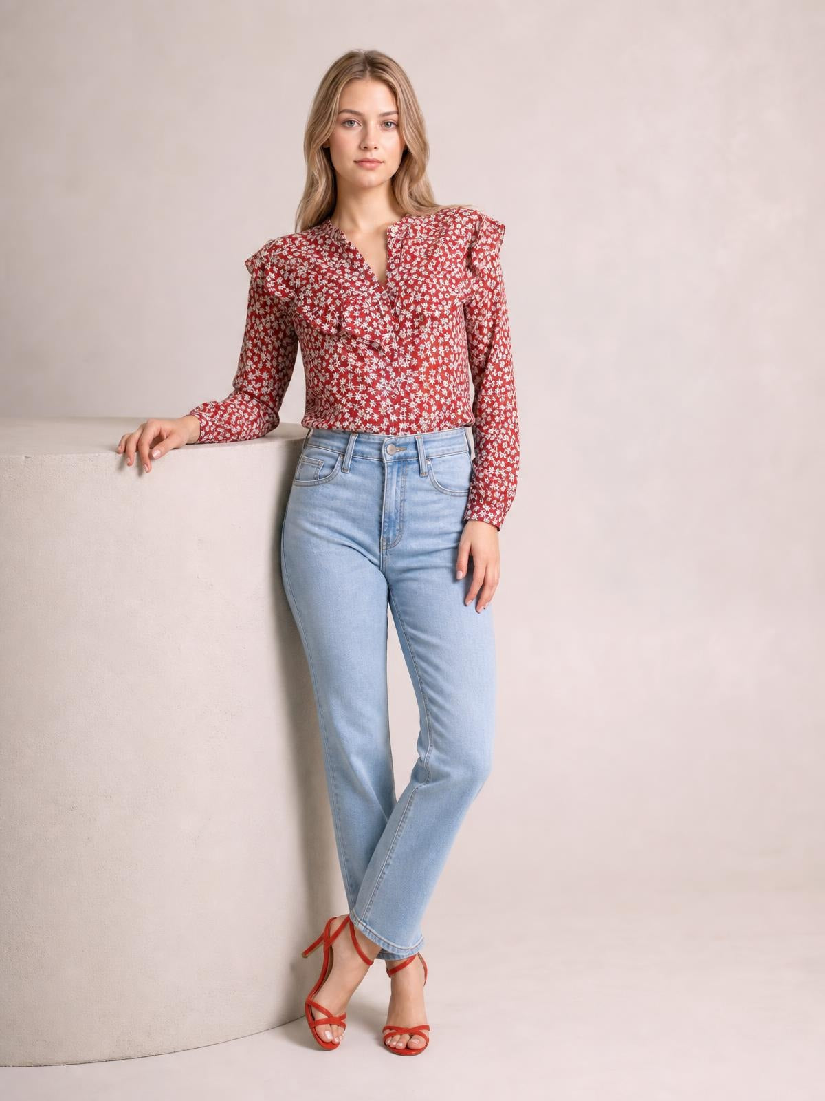 A model poses with crossed ankles in a red floral printed blouse featuring a statement ruffled collar, button front, long sleeves, and a softly fitted silhouette.