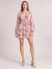 Model stands in a pink printed surplice mini dress with a V-neckline, tie waist, long balloon sleeves, flared silhouette, and tiered ruffle hem.
