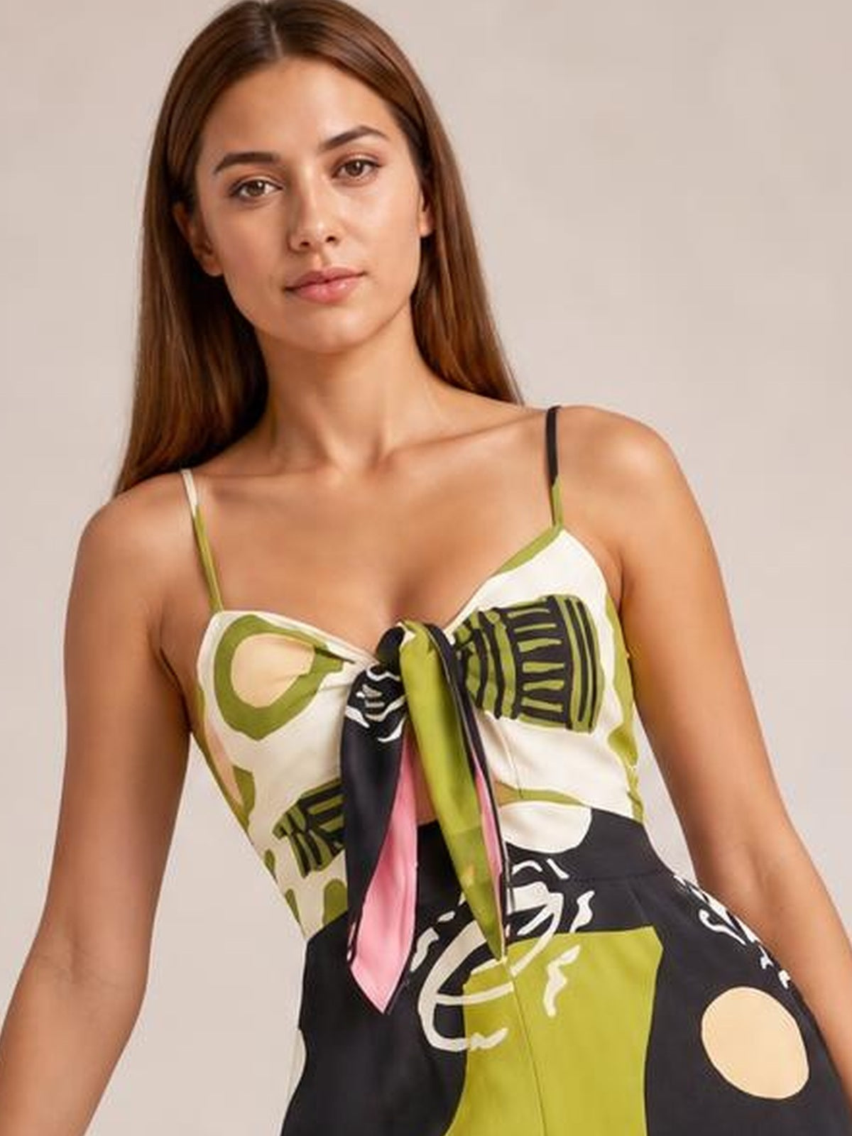 Model poses in a printed jumpsuit with thin straps, a sweetheart neckline, tied front, bold olive, black, white, and pink pattern, and a relaxed silhouette.