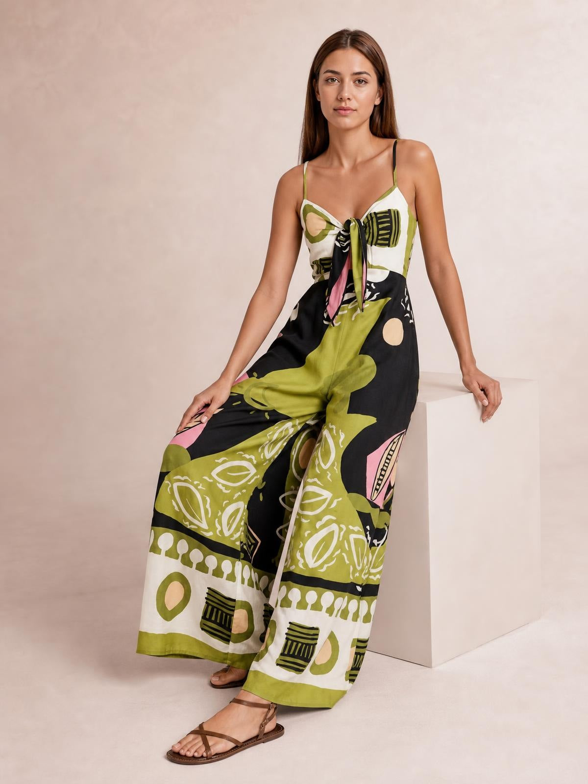 Model in a printed sweetheart-neck jumpsuit with tie-front bodice, slim straps, fitted waist, and flowing wide legs in black, olive, white, and pink.