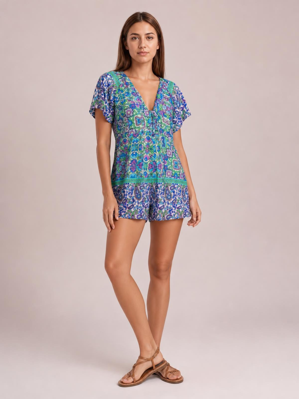 A model stands with crossed legs in a colorful printed tie-front romper featuring short sleeves, a V-neckline, and a relaxed shorts silhouette.