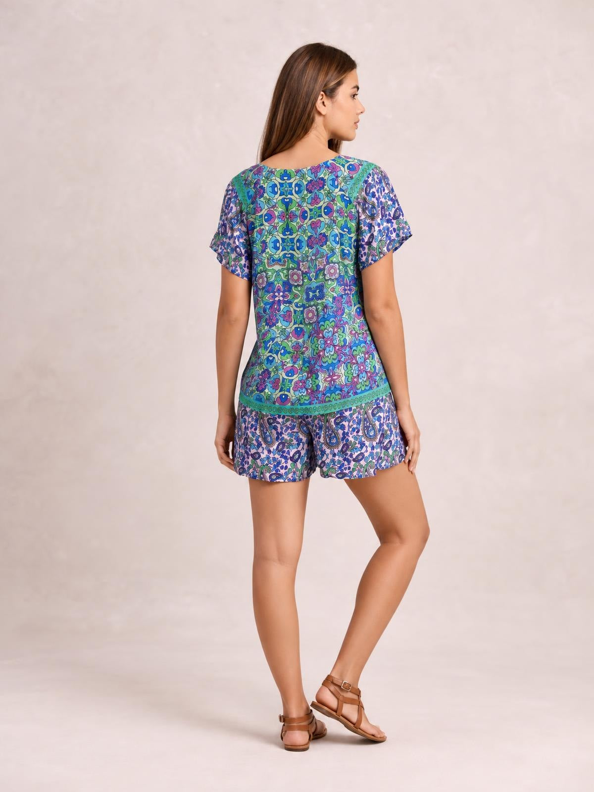 A model poses from the back in a colorful blue, green, and purple printed short-sleeve romper with a relaxed top and loose shorts silhouette.