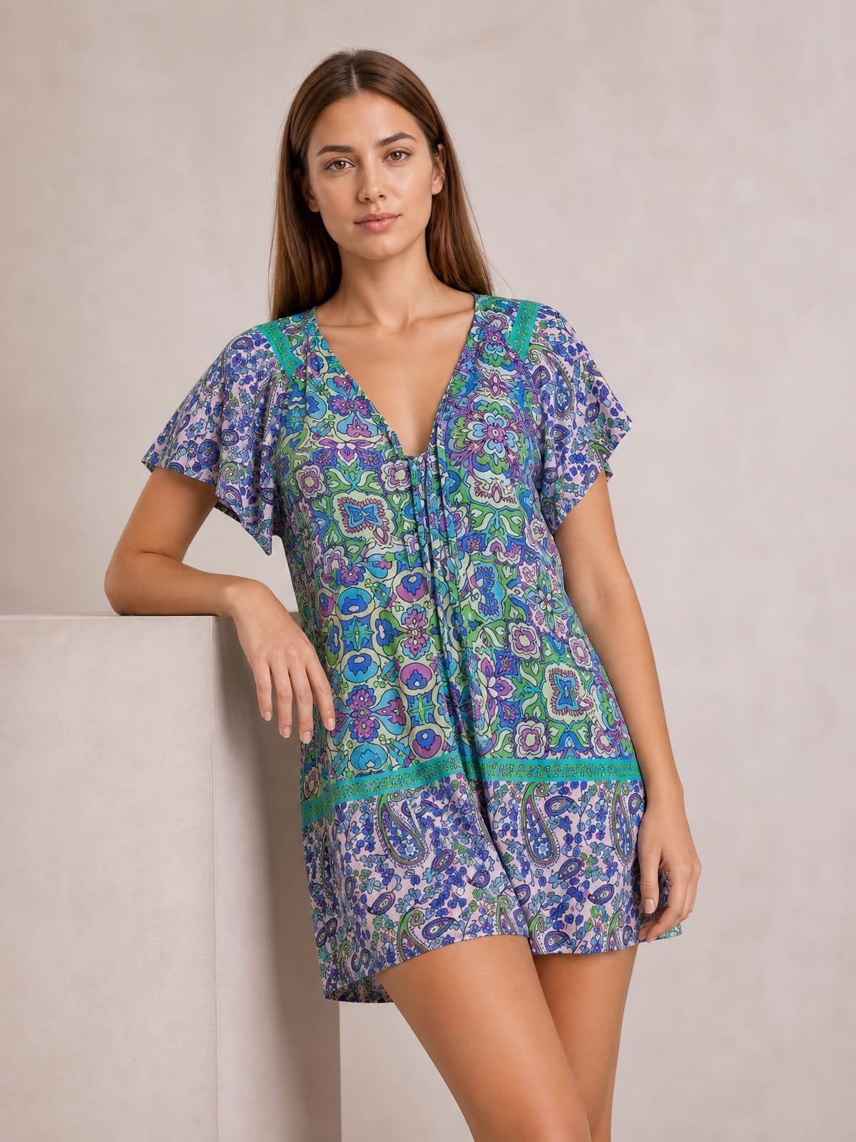 Model wears a colorful printed tie-front romper with short flutter sleeves, a V-neckline, relaxed silhouette, and floral paisley pattern while leaning casually.