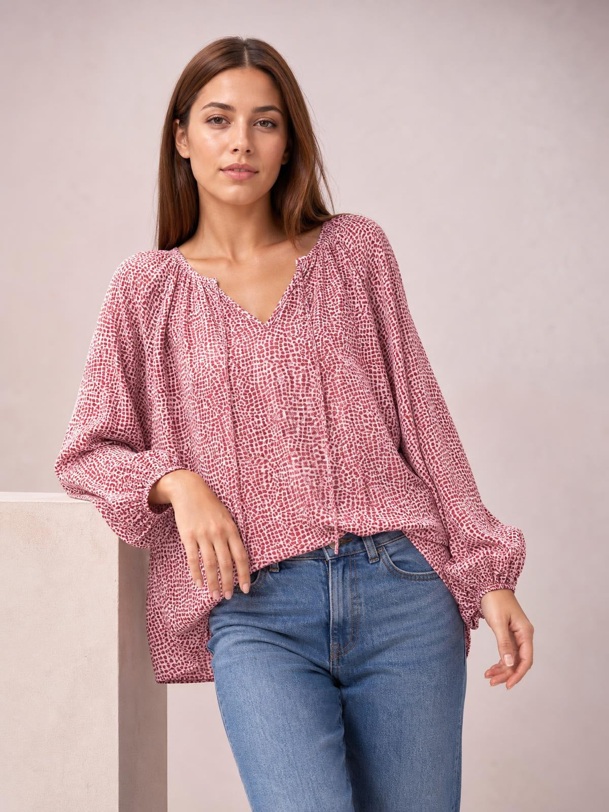 A pink and white printed blouse features a gathered V-neckline, long balloon sleeves with cuffs, and a relaxed long silhouette as the model poses with one hand at her hip.