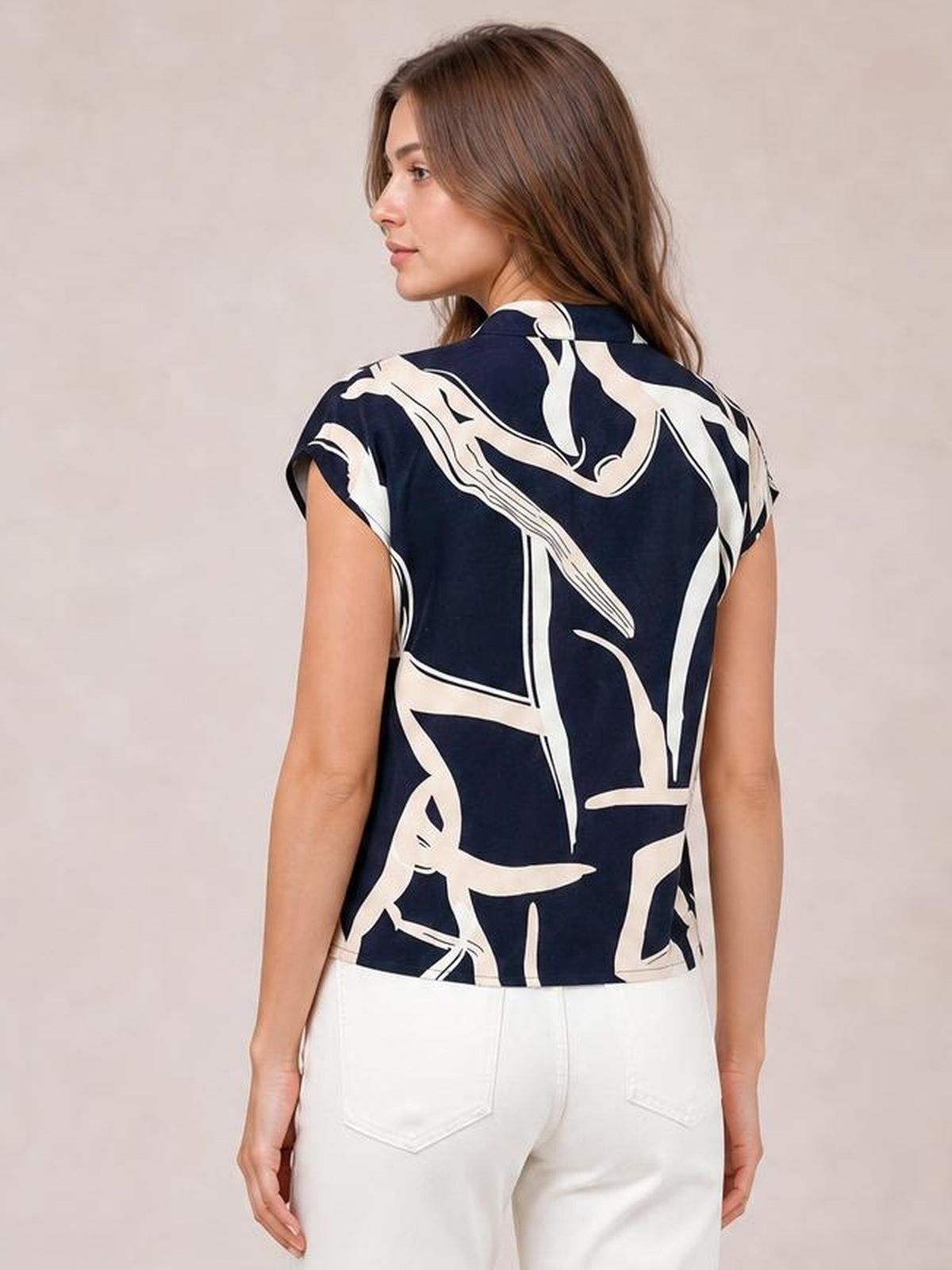 A model stands facing away in a navy printed cap sleeve blouse with an abstract cream pattern, high neckline, and relaxed straight silhouette.