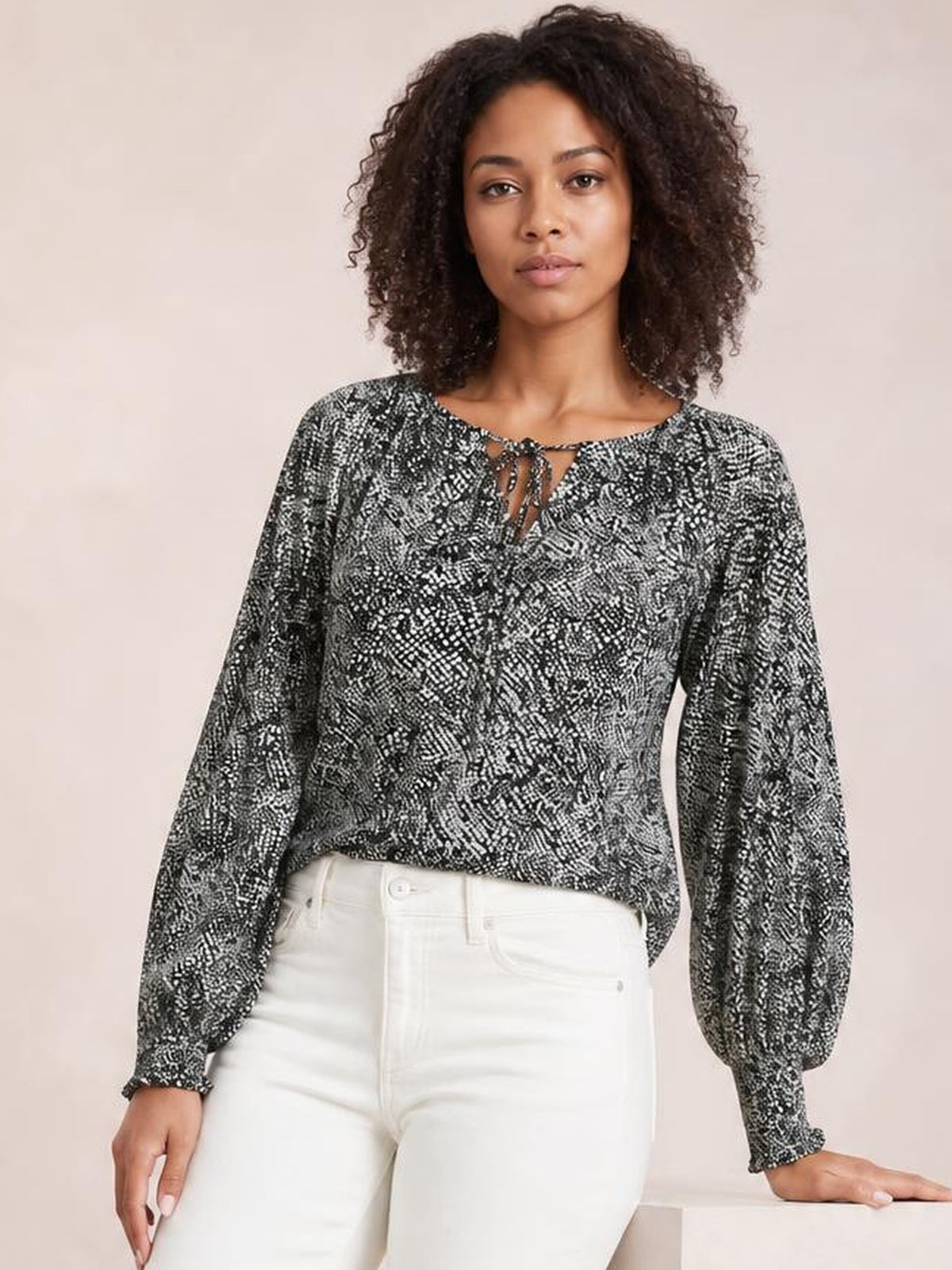Model wearing a black-and-white printed tie neck blouse with billowy lantern sleeves, gathered cuffs, and a relaxed silhouette, standing with one hand on a ledge.