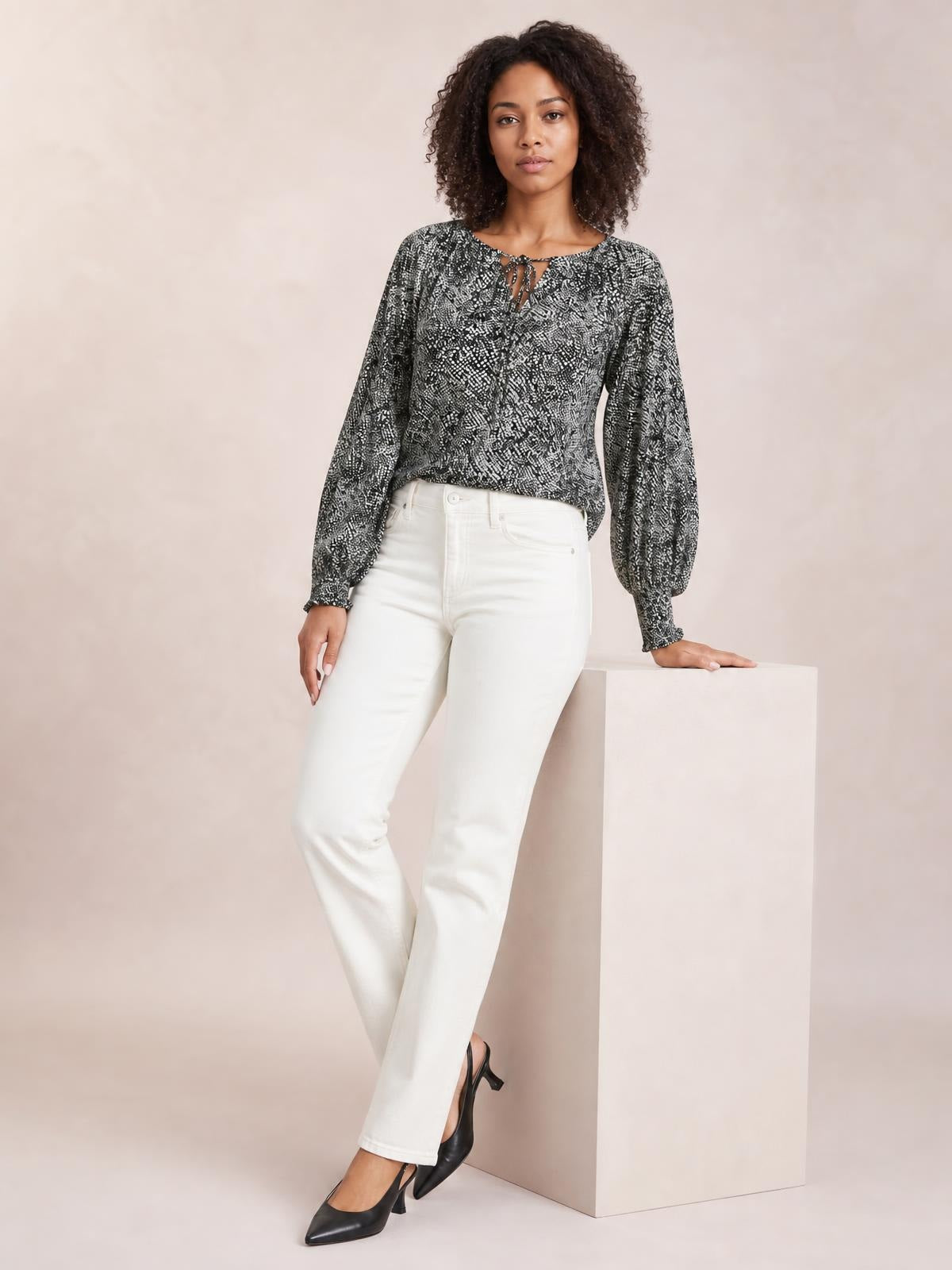 Model in a black and white printed blouse with a tied neckline, relaxed silhouette, and voluminous lantern sleeves, leaning against a pedestal.
