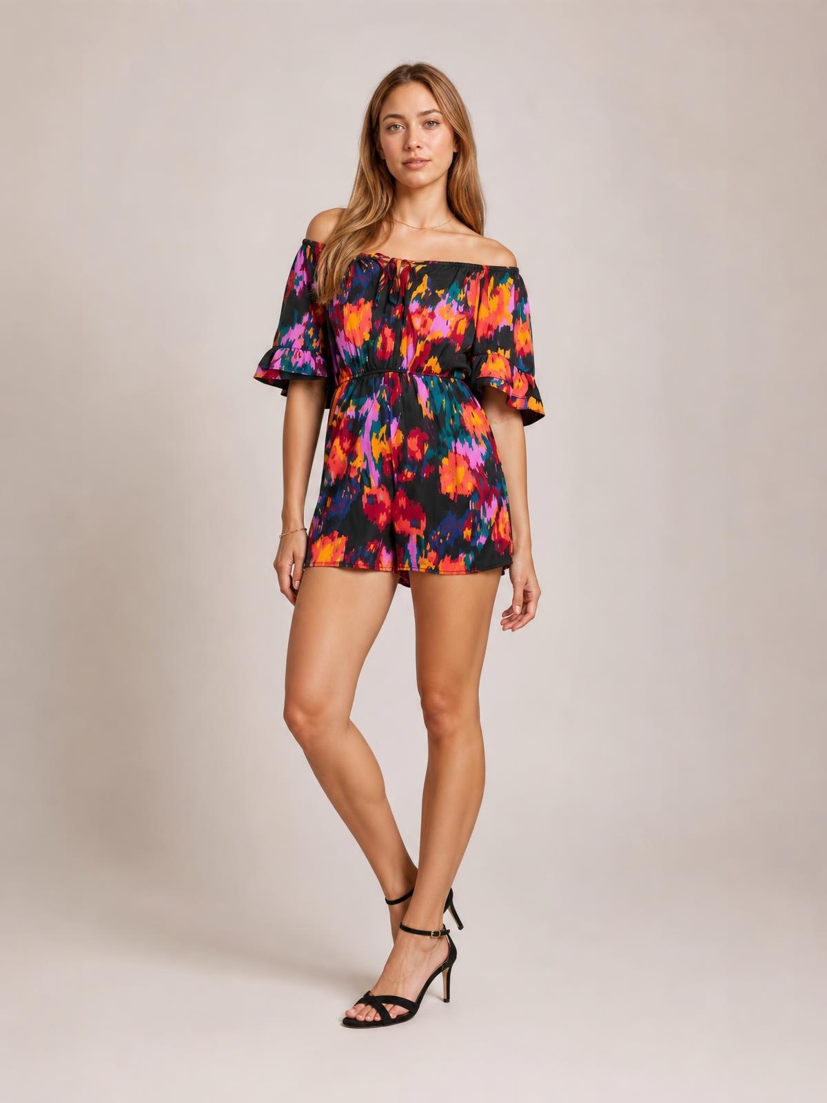 A model poses with crossed legs in a multicolored printed romper featuring a boat neckline, short flounce sleeves, a drawstring waist, and short legs.