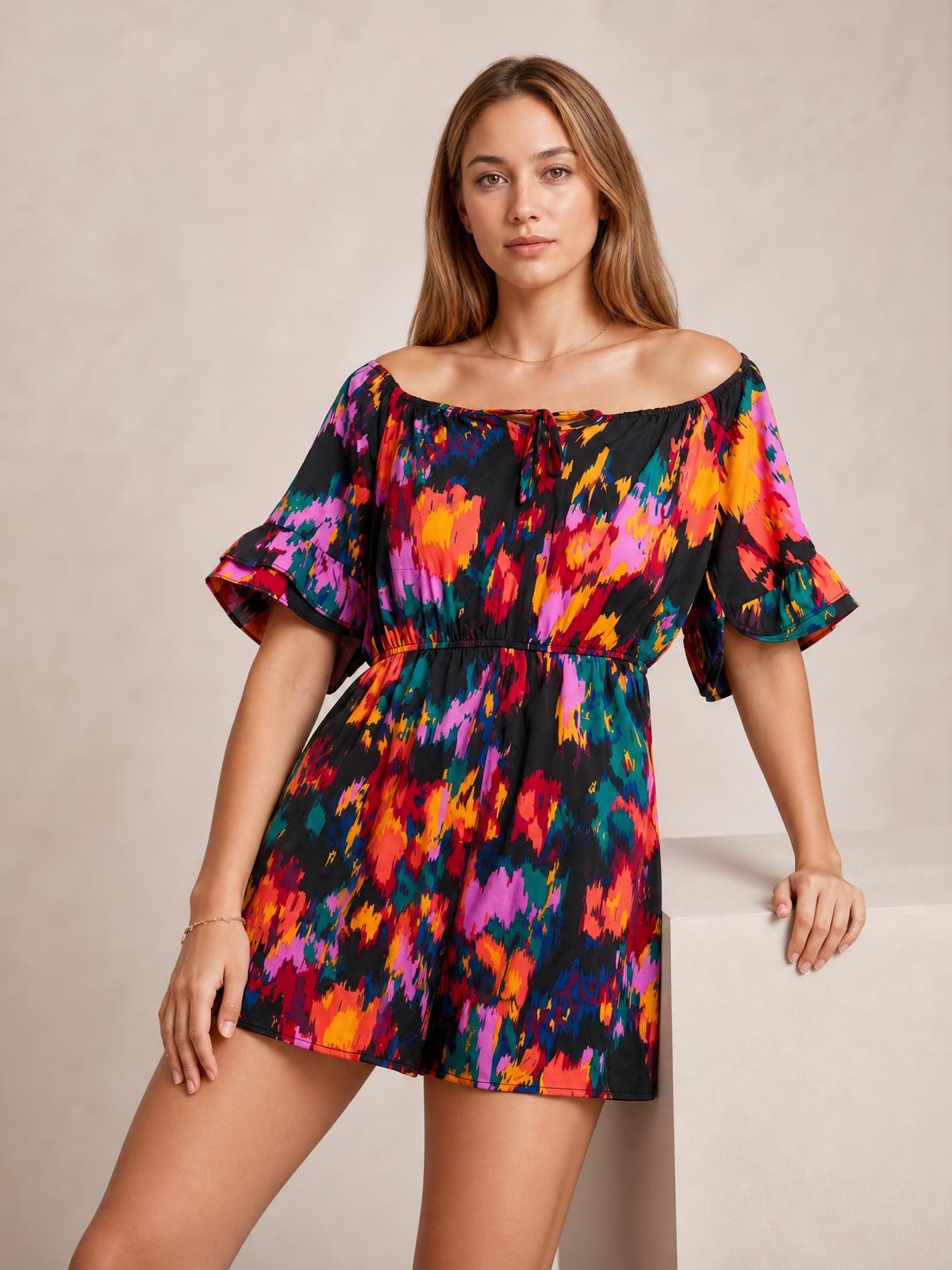 A model poses in a multicolored printed romper with a boat neckline, tied drawstring waist, short flounce sleeves, and relaxed short-leg silhouette.
