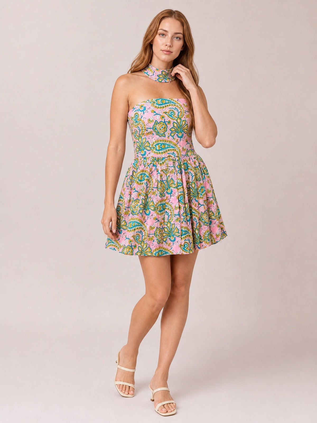 Model wears a pink paisley-printed tube mini dress with a fitted smocked bodice, gathered waist, softly flared skirt, and halter neck detail, posing with crossed legs.