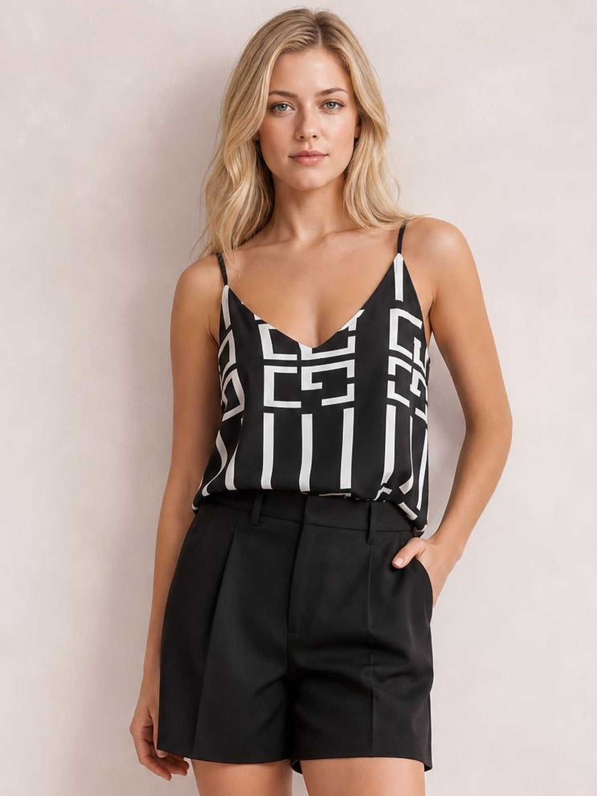 Model wearing a black printed V-neck cami with thin straps and a relaxed silhouette, paired with black shorts while standing with one hand in a pocket.