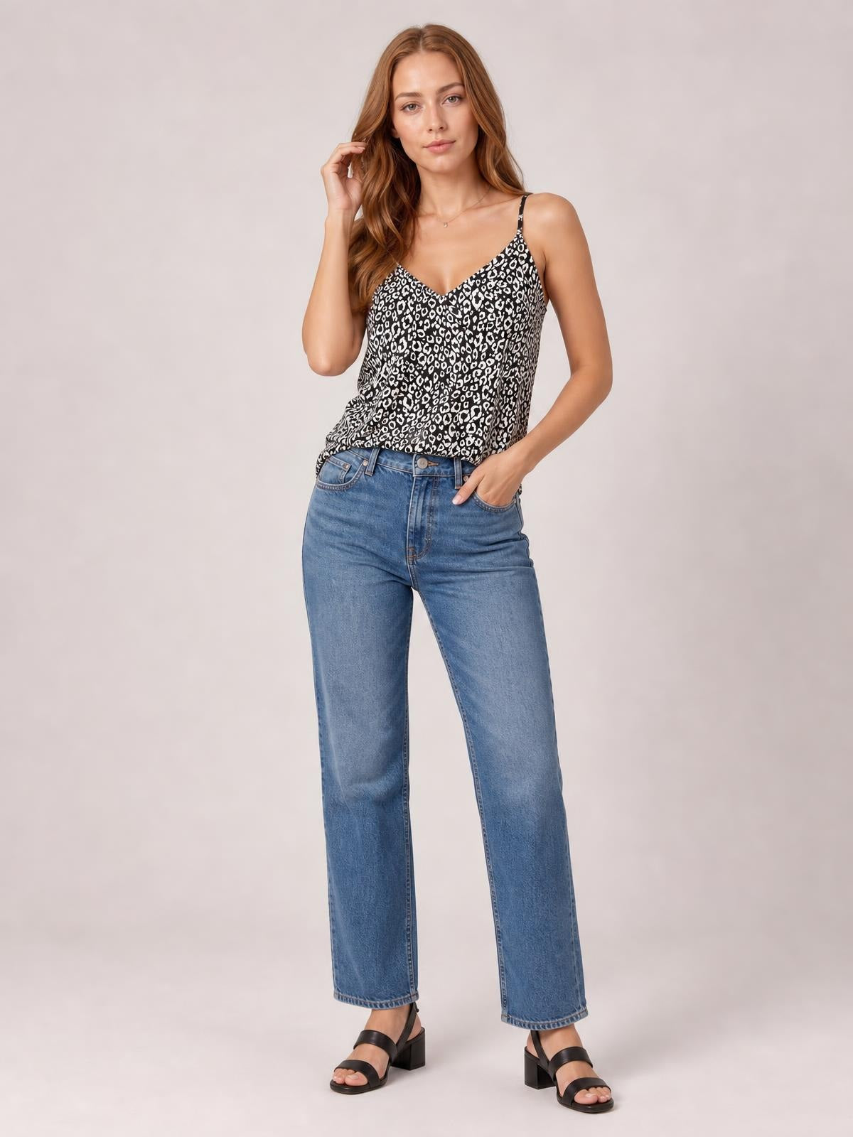 Model wearing a black-and-white printed V-neck cami with thin straps and a relaxed silhouette, paired with blue wide-leg jeans while posing with one hand in a pocket.