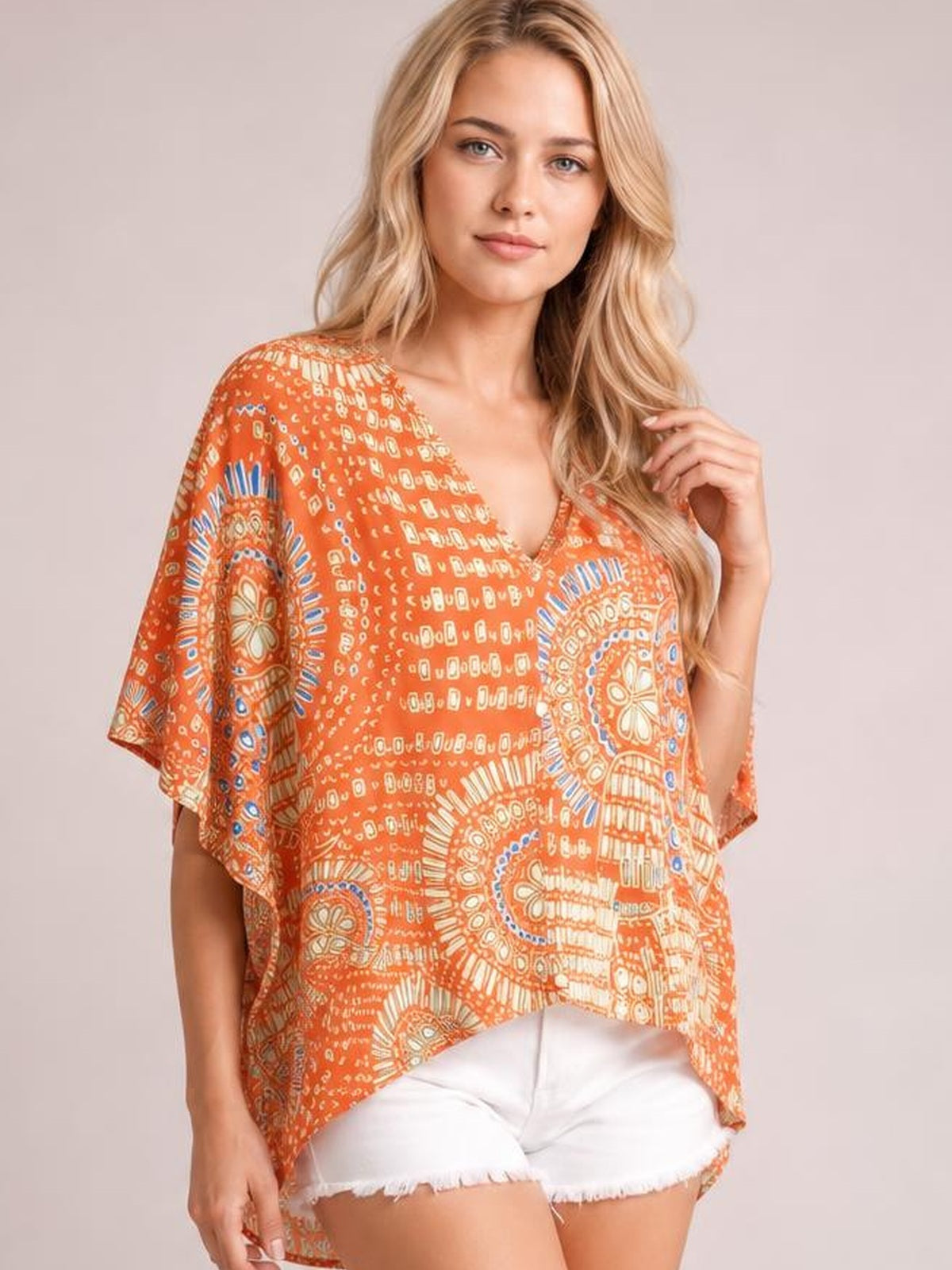 A model poses in an orange printed V-neck top with loose, flowing silhouette, wide half sleeves, and an asymmetrical draped hem while touching her hair.