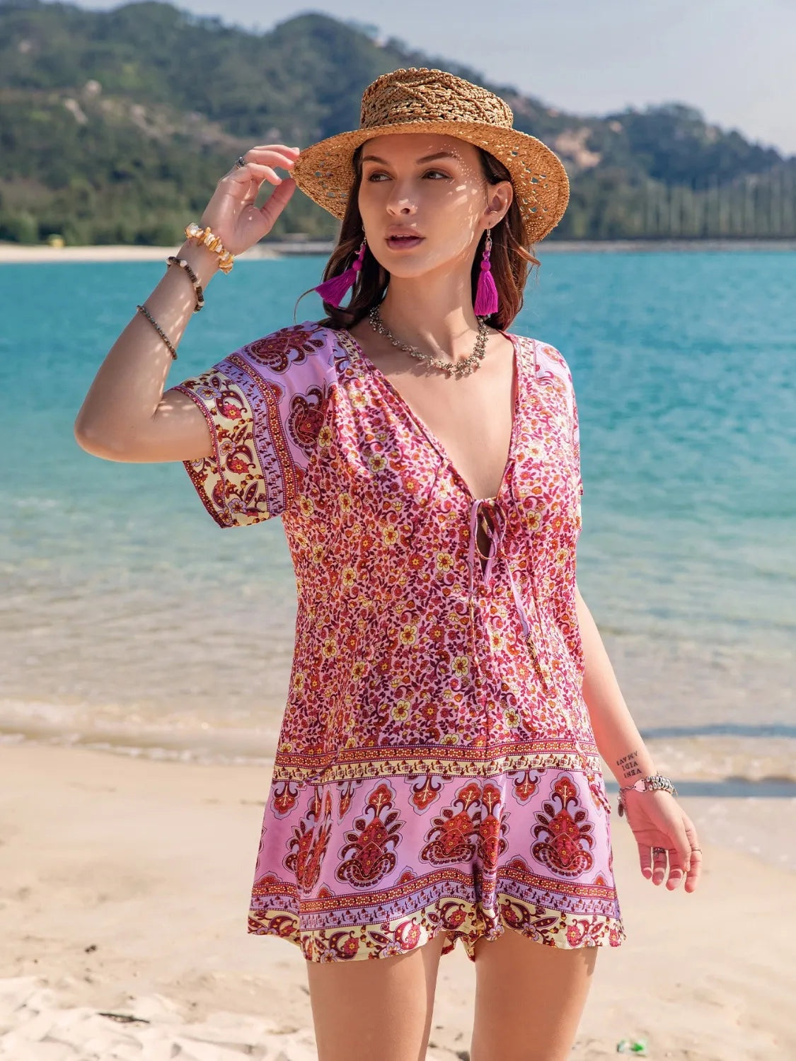 Model poses in a pink and burgundy printed V-neck short-sleeve romper with a tied waist, relaxed silhouette, and flared shorts.