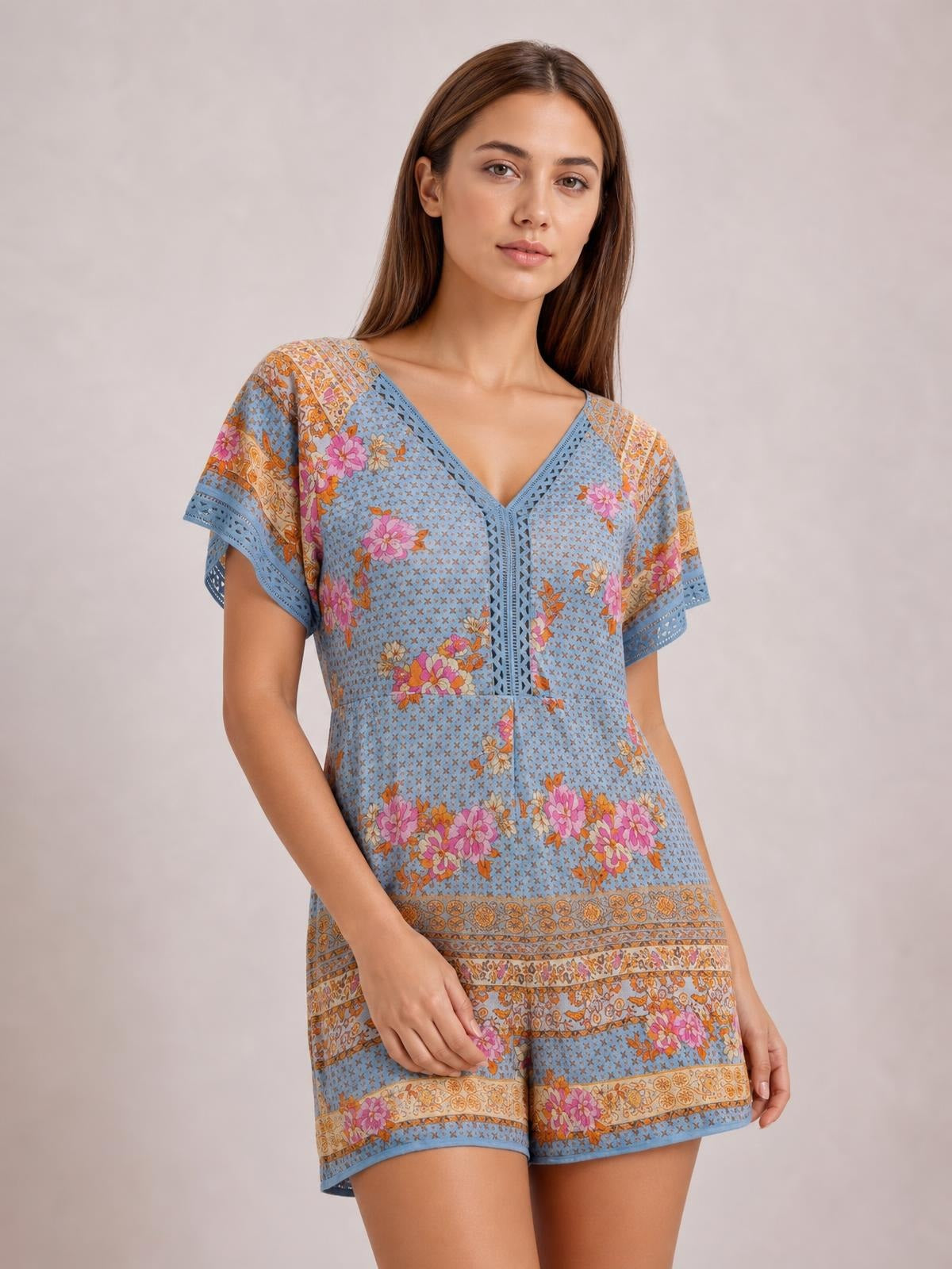Model poses in a blue floral printed romper with a V-neck, short flutter sleeves, semi-sheer fabric, defined waist, and relaxed shorts silhouette.